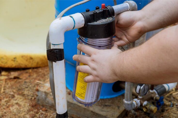 Hands installing a clear water filter. The filter is attached to white PVC pipes and a blue tank.