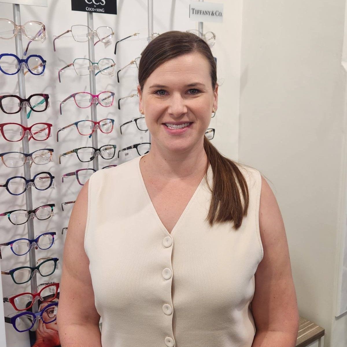Woman with glasses smiles in front of a sign with blue circles. She wears a maroon cardigan.