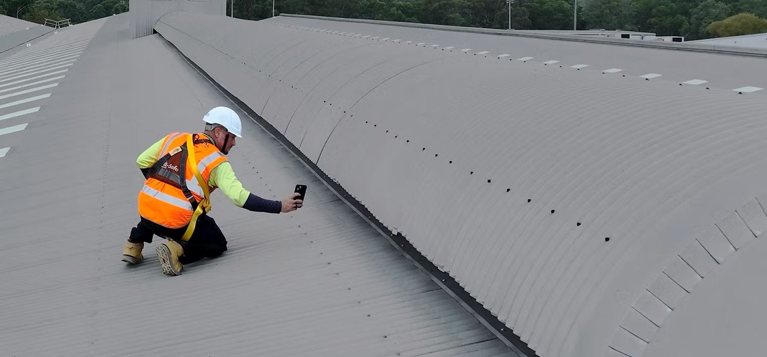 A person wearing a high-visibility vest and safety helmet kneels on a corrugated metal roof, taking a photo of a seam.