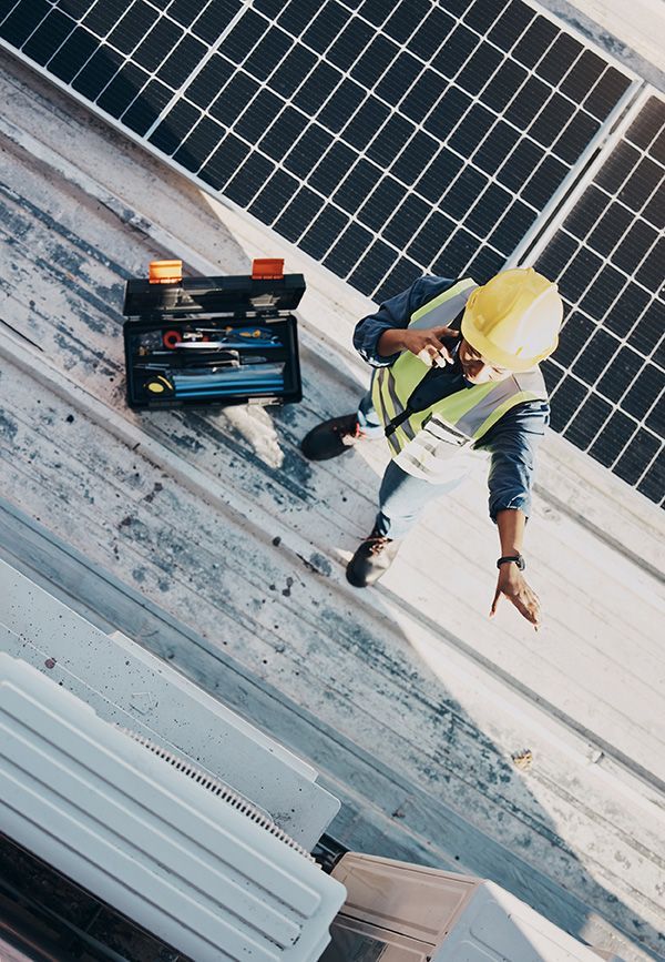 A worker in a high-visibility vest and hard hat talks on a phone while standing on a roof with solar panels and a toolbox.