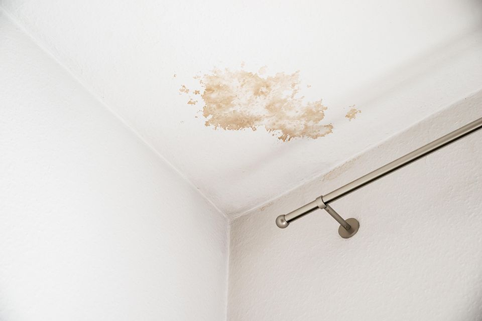 A light-brown water stain on a white ceiling corner, located above a metal curtain rod mounted to the wall.