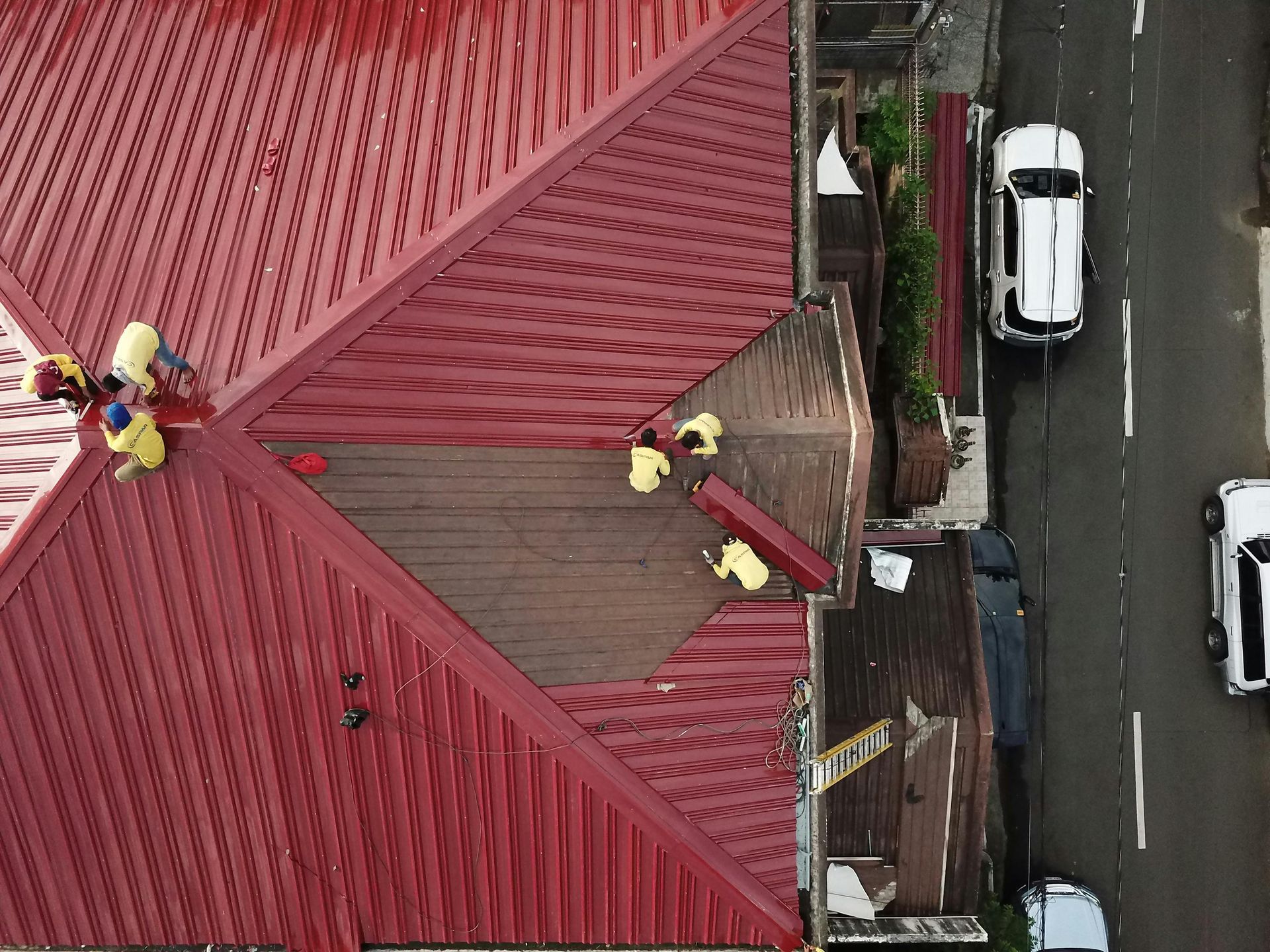 Aerial view of workers in yellow shirts installing a red metal roof on a building near a street with two cars.