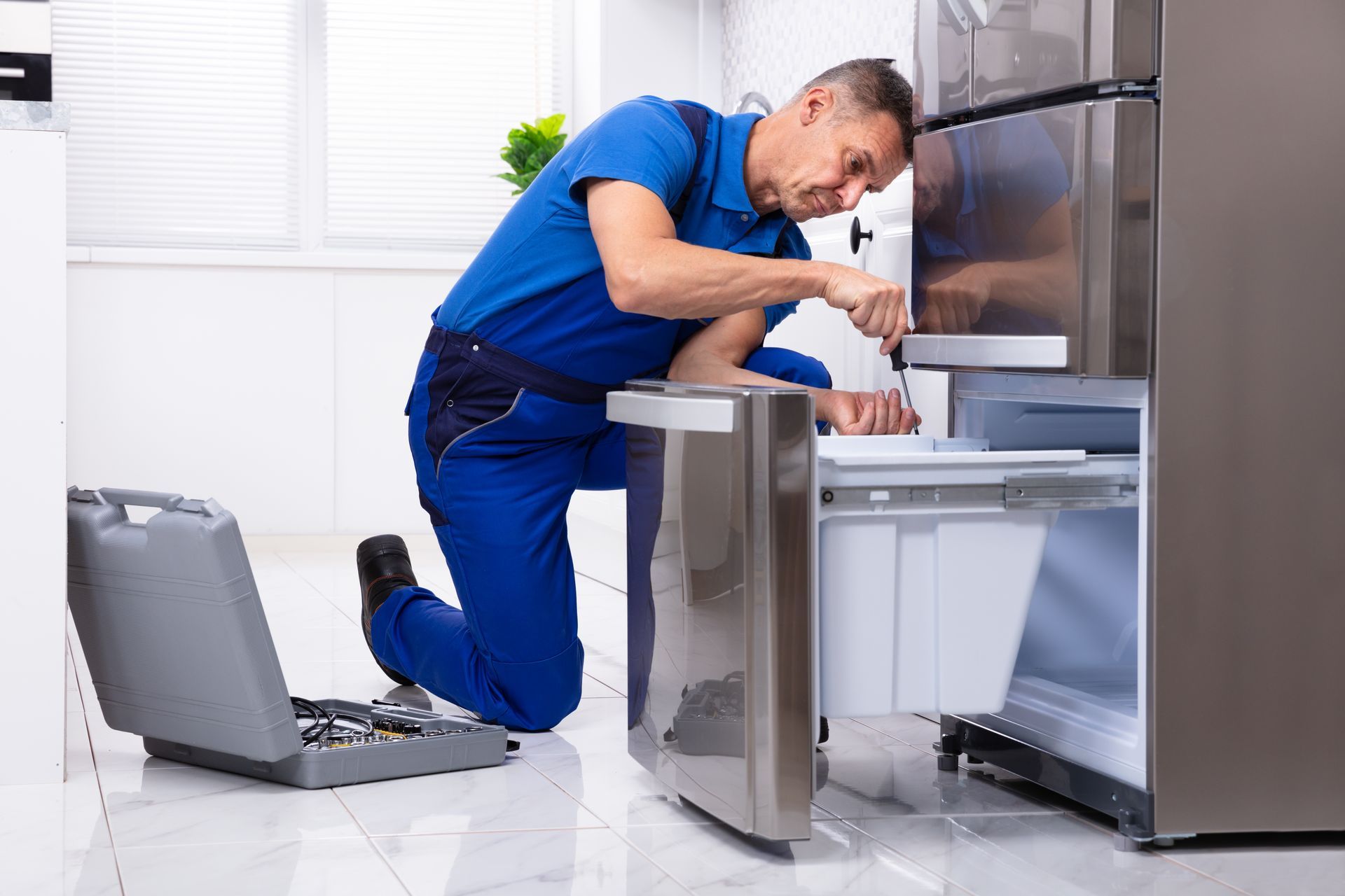 A repair technician in blue overalls fixes a refrigerator with a toolbox on the floor.