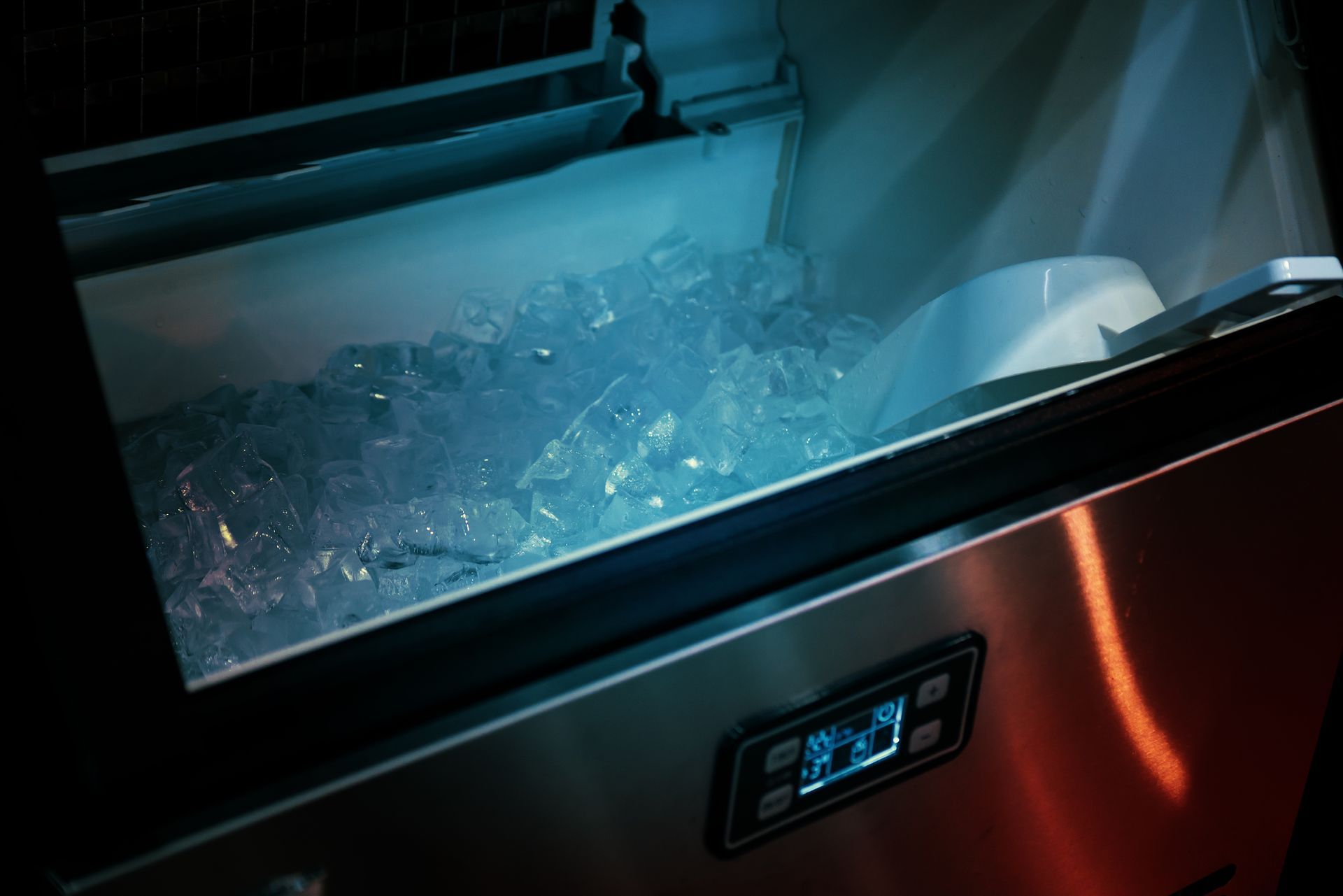 Close-up of an ice machine with a bin full of ice cubes, lit by cool blue light, and a control panel.