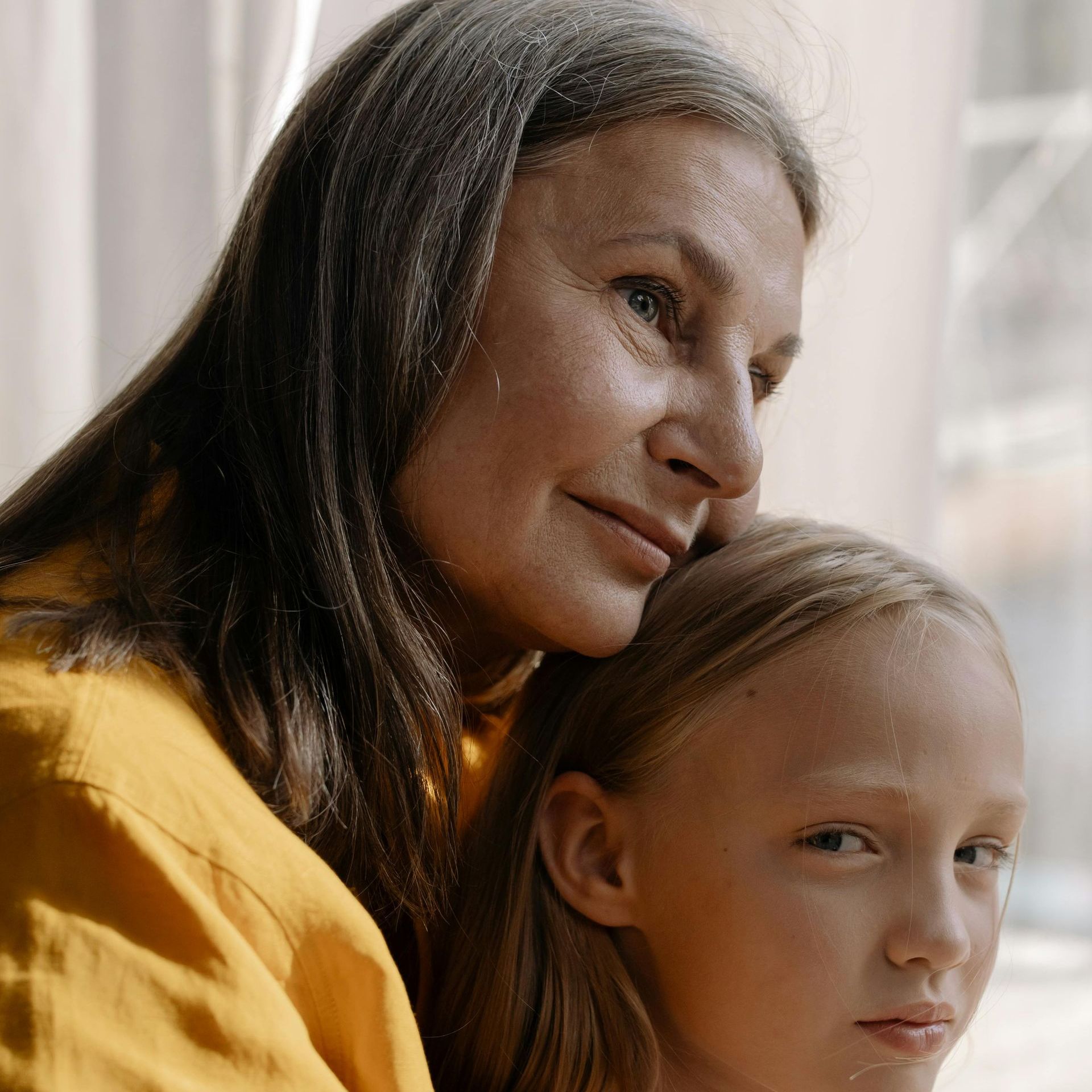 A woman in a yellow shirt is hugging a little girl in front of a window.