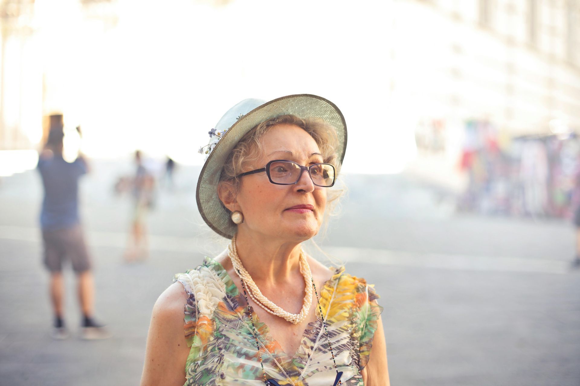 An elderly woman wearing a hat and glasses is standing on a street.