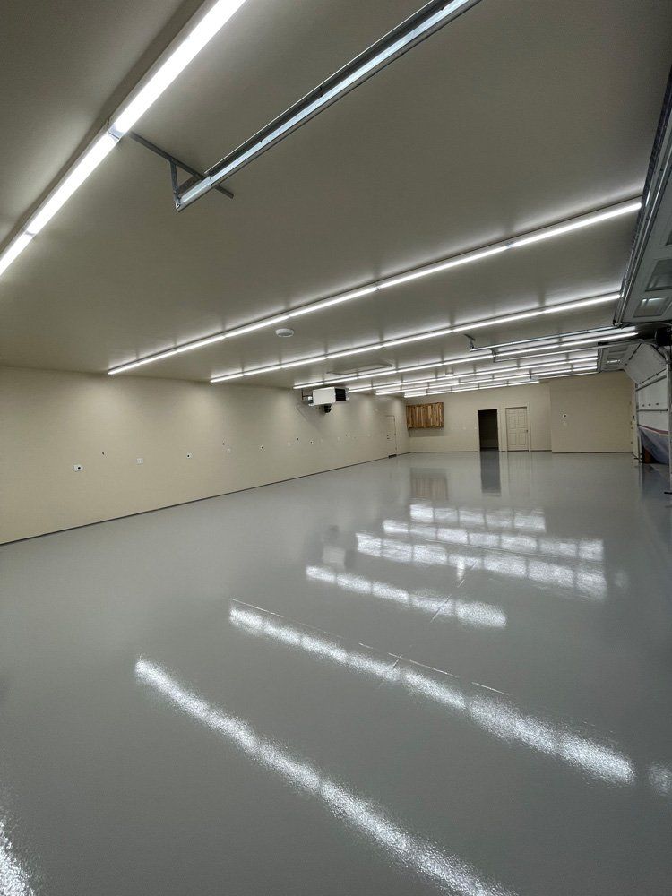 Empty, light-filled workshop or warehouse. Shiny gray floor, white walls and ceiling. Fluorescent lights reflect.