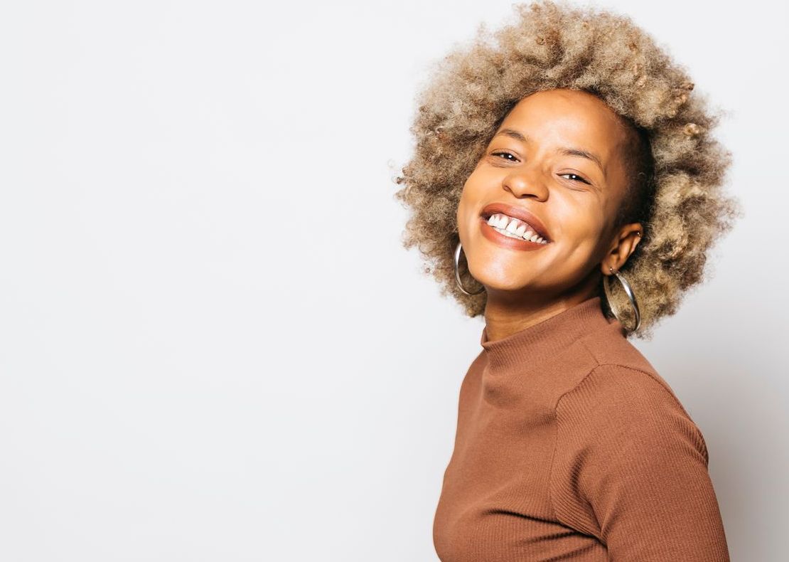 A woman with curly hair is smiling and wearing a brown sweater.