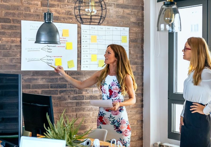 Two women are standing in front of a whiteboard in an office.