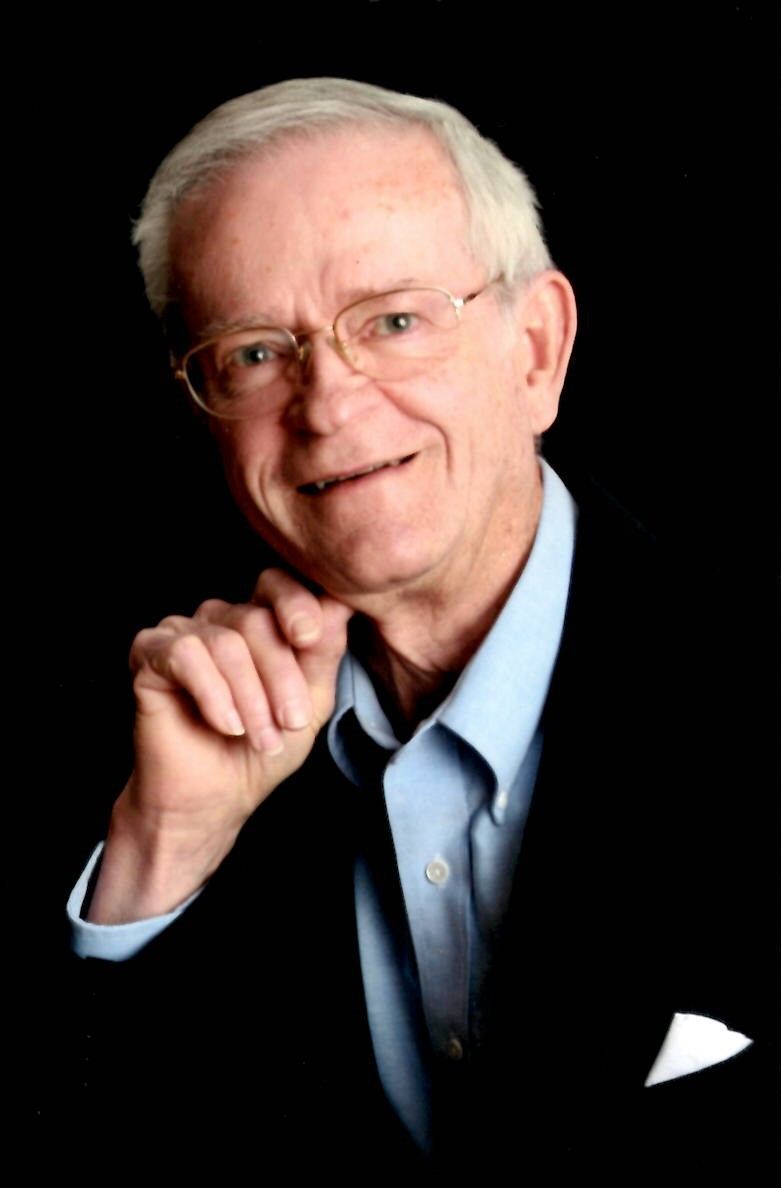 A man wearing glasses and a blue shirt is smiling with his hand on his chin.