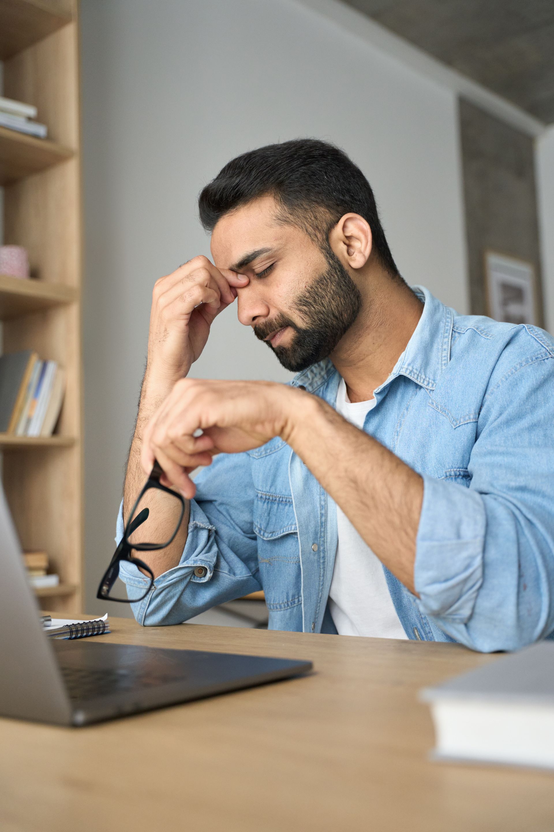 A man is sitting at a desk with a laptop and holding his head in frustration.