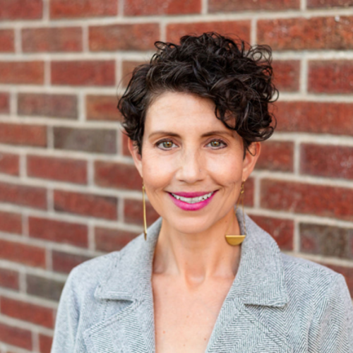 A woman with curly hair is smiling in front of a brick wall.