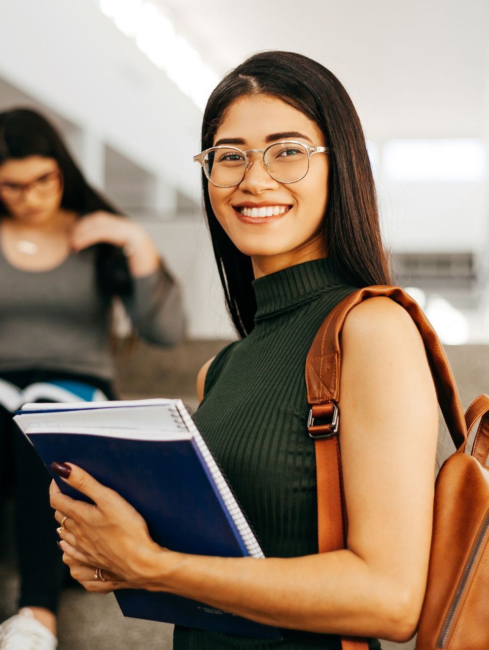 A woman with glasses is holding a book and a backpack.