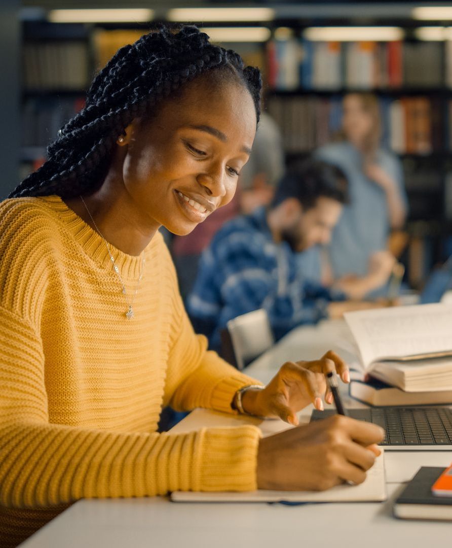 A woman in a yellow sweater is sitting at a desk in a library.