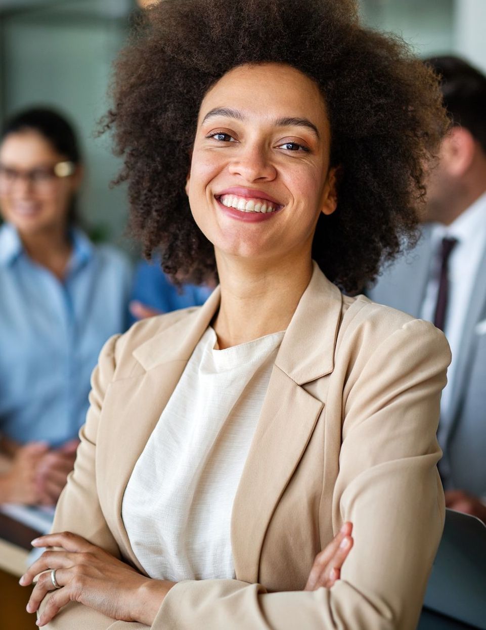 A woman with curly hair is smiling with her arms crossed in front of a group of people.