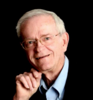 A man wearing glasses and a blue shirt is smiling with his hand on his chin.