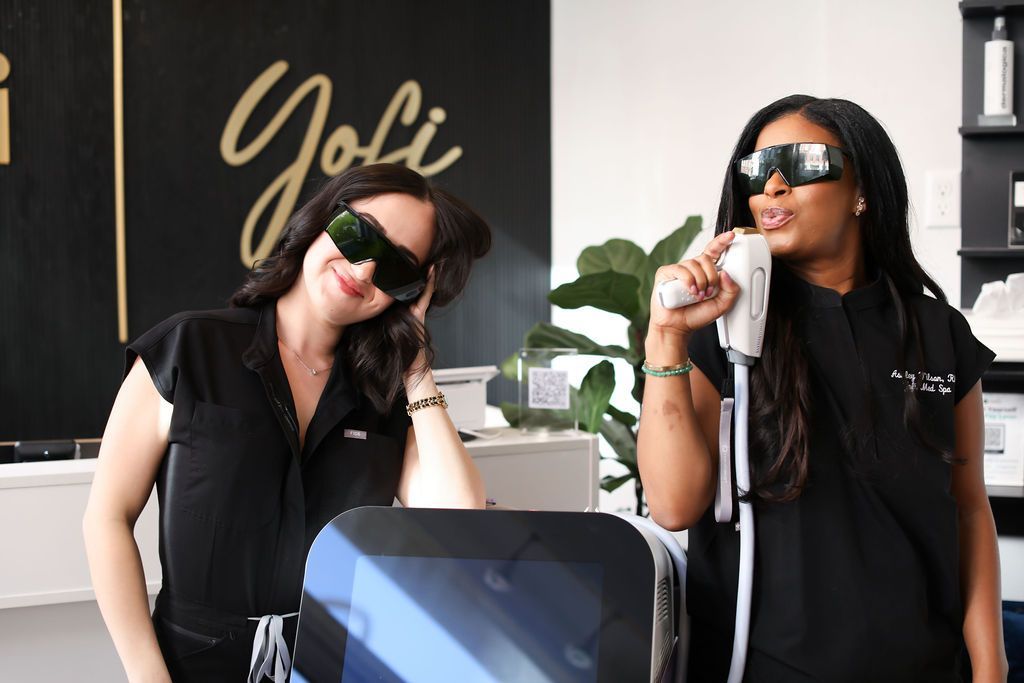 Two women wearing sunglasses are standing next to each other in front of a sign that says joli