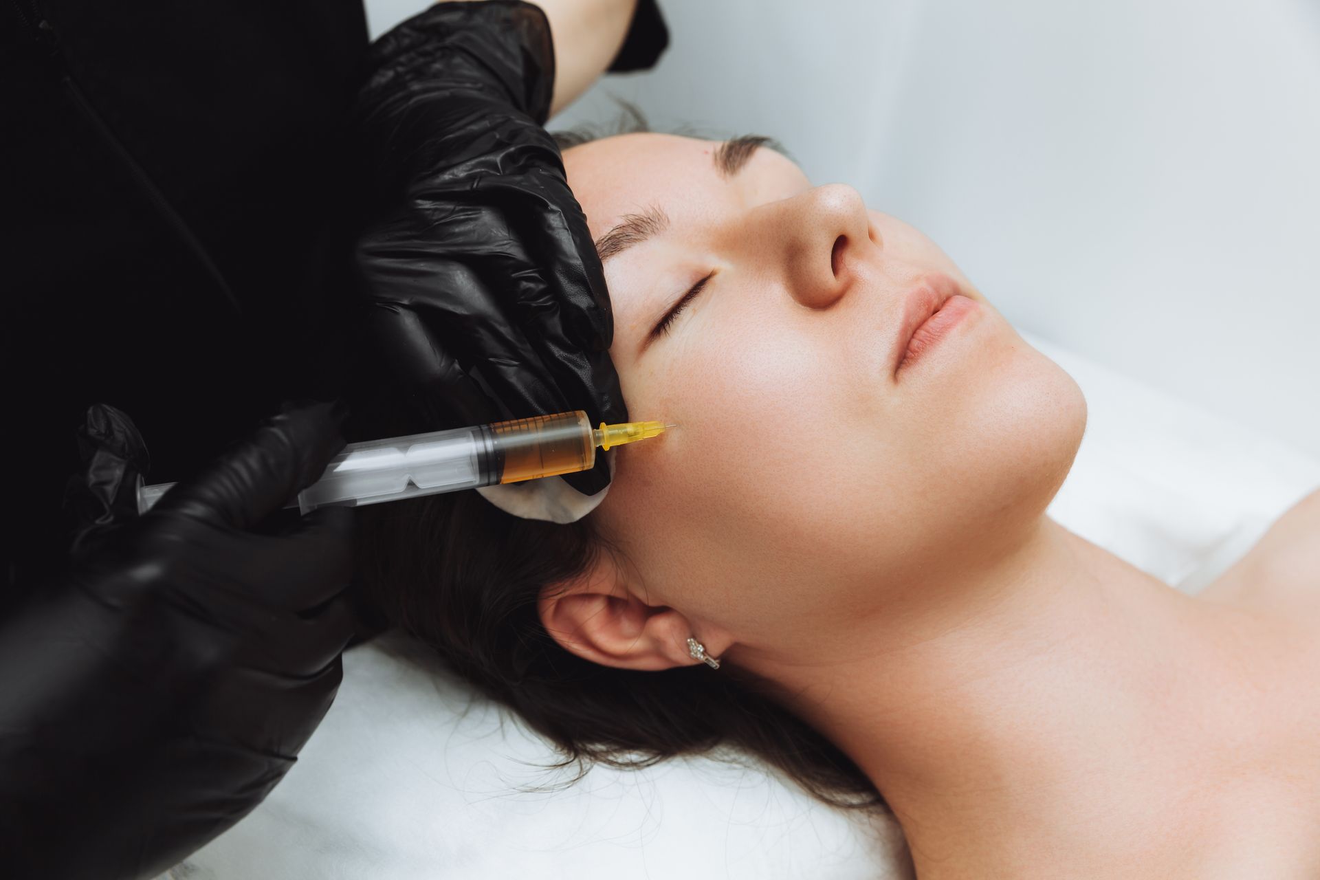 Person receiving facial injection; gloved hand holds syringe near cheek.