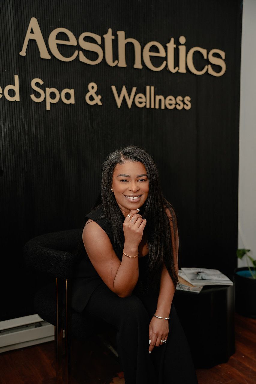 Woman seated in front of “Aesthetics Med Spa & Wellness” sign. She smiles, touches her chin. Dark wall.