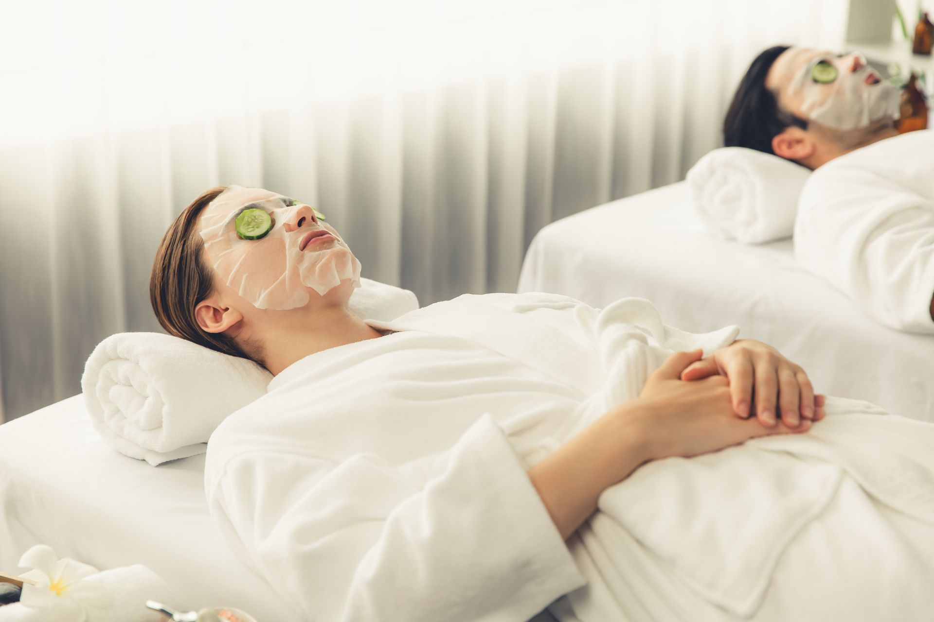 Two people in white robes relaxing on massage tables with face masks and cucumber slices on their eyes in a spa.