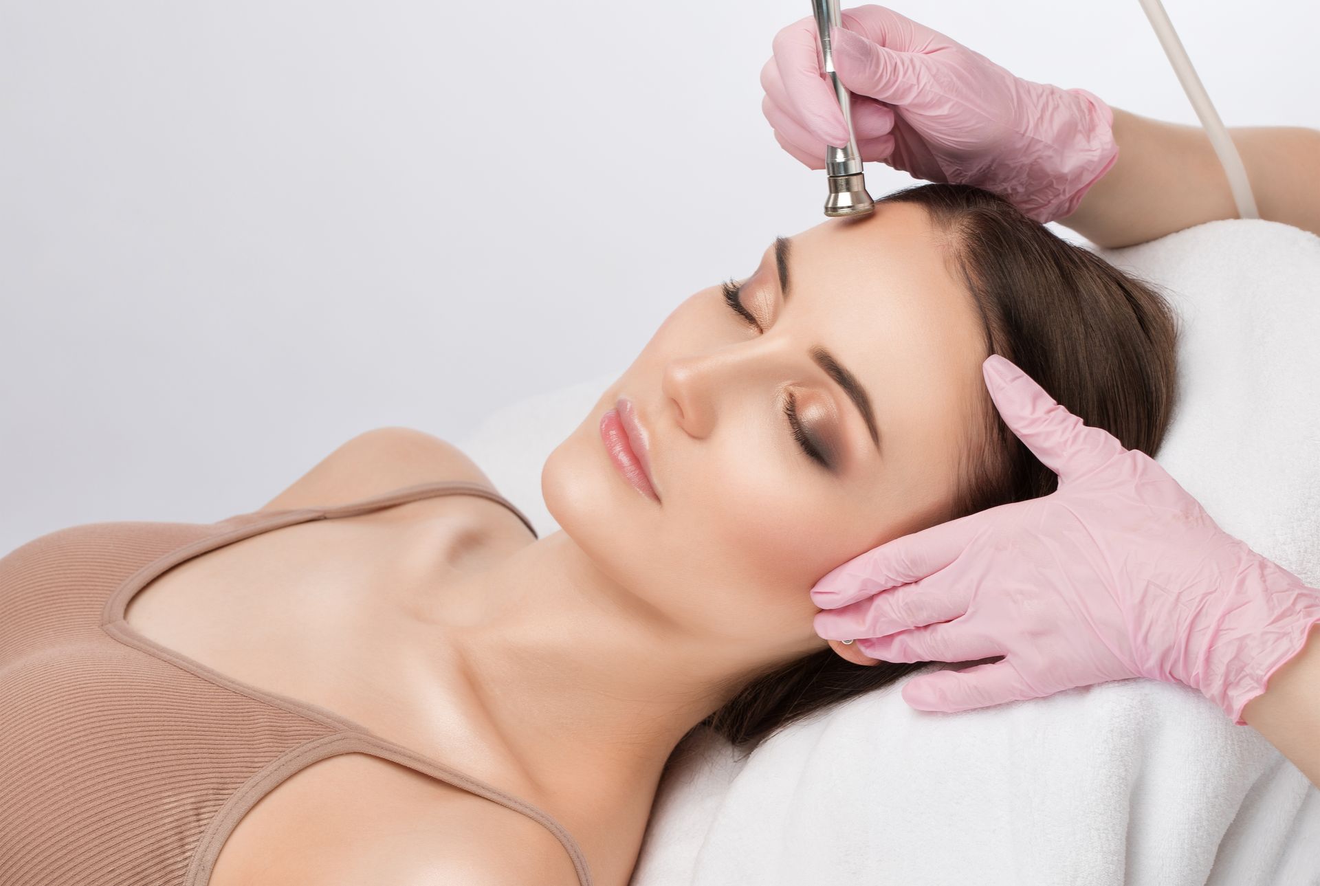 Woman receiving facial treatment, lying on white towel, hands in pink gloves, tool on forehead.