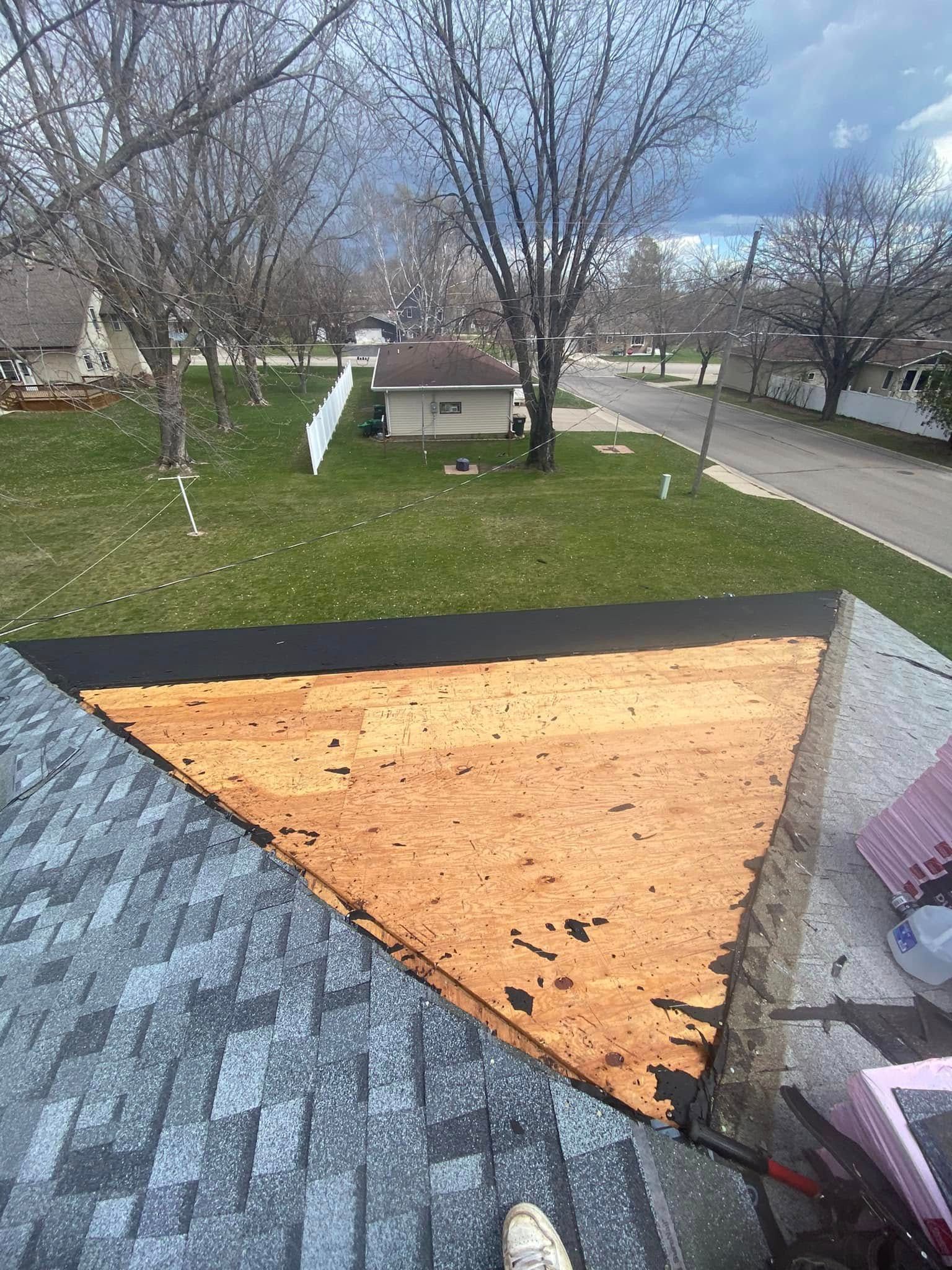 Damaged roof with exposed wood sheathing after shingle removal, viewed over a suburban yard.