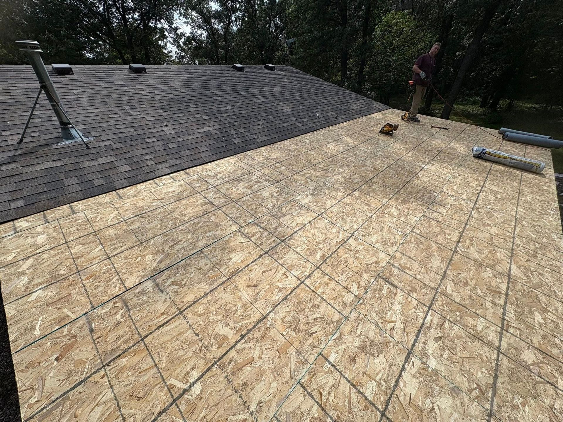 Roof under construction with exposed plywood, dark shingles, and two workers in the background