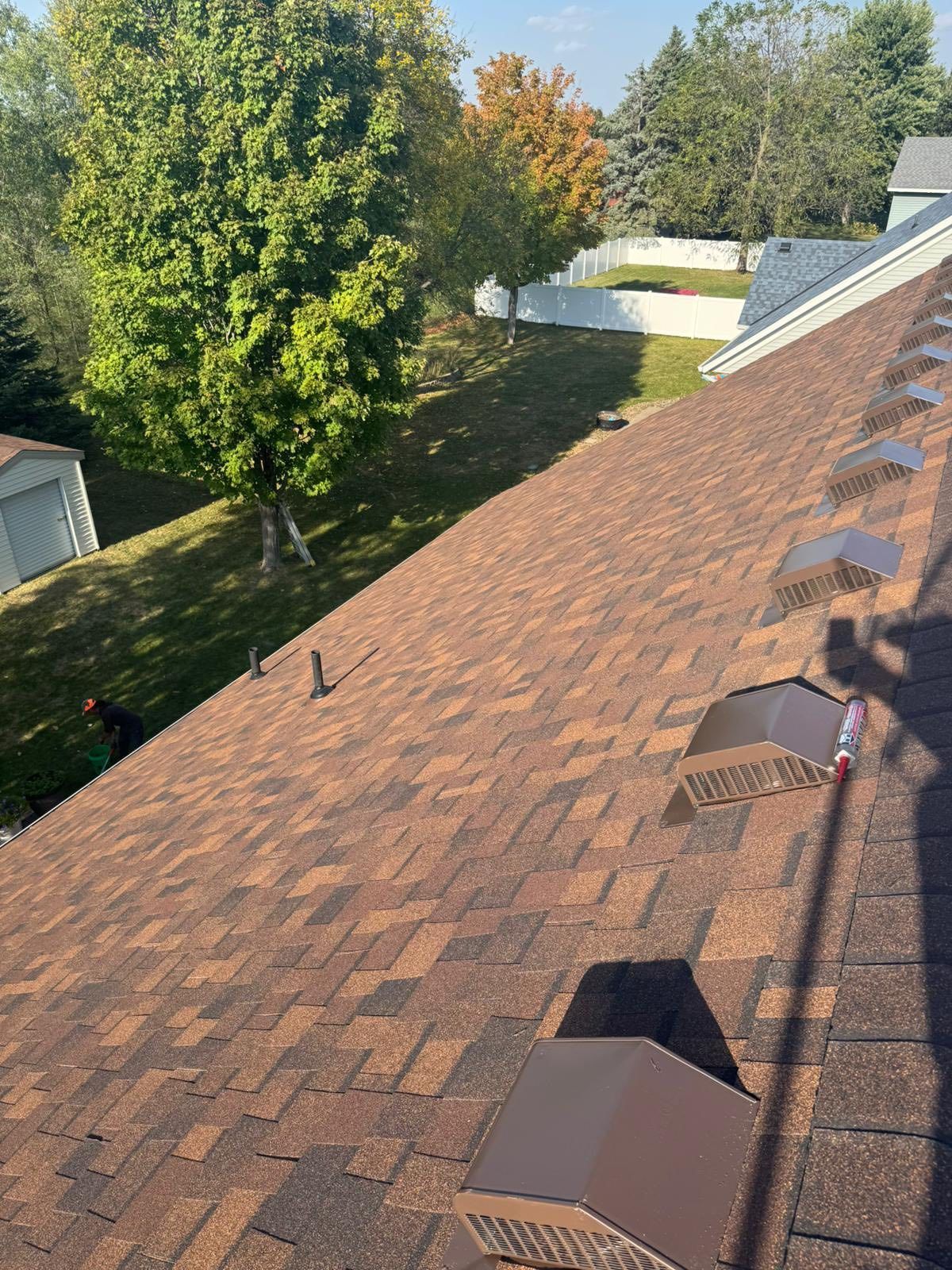 Rooftop with brown shingles and several vents, overlooking trees and houses in a suburban neighborhood
