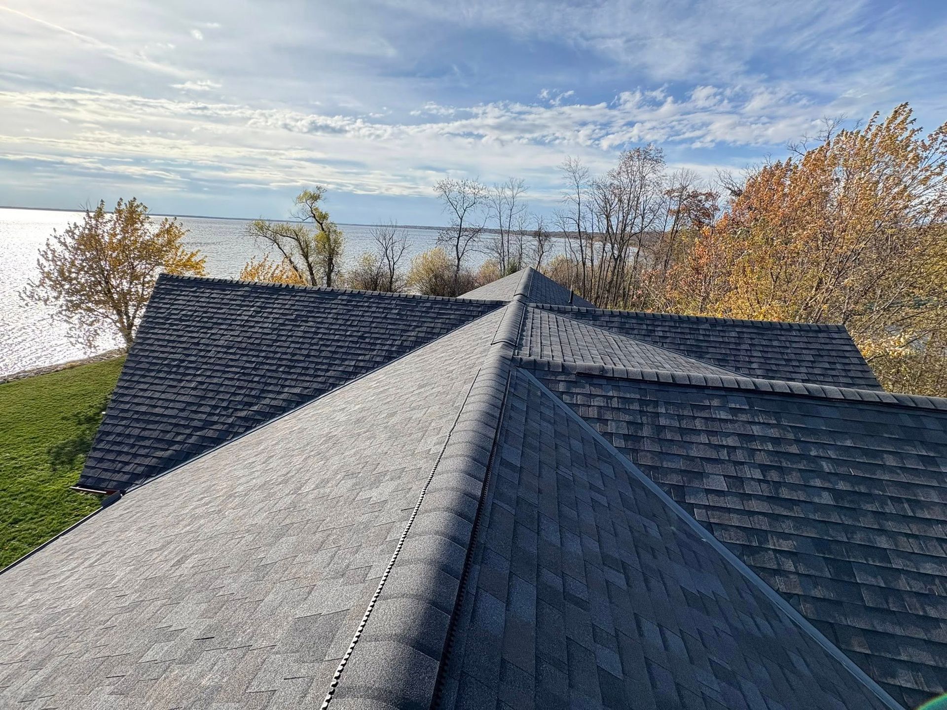 Dark shingled rooftop overlooking a lake with autumn trees under a partly cloudy sky