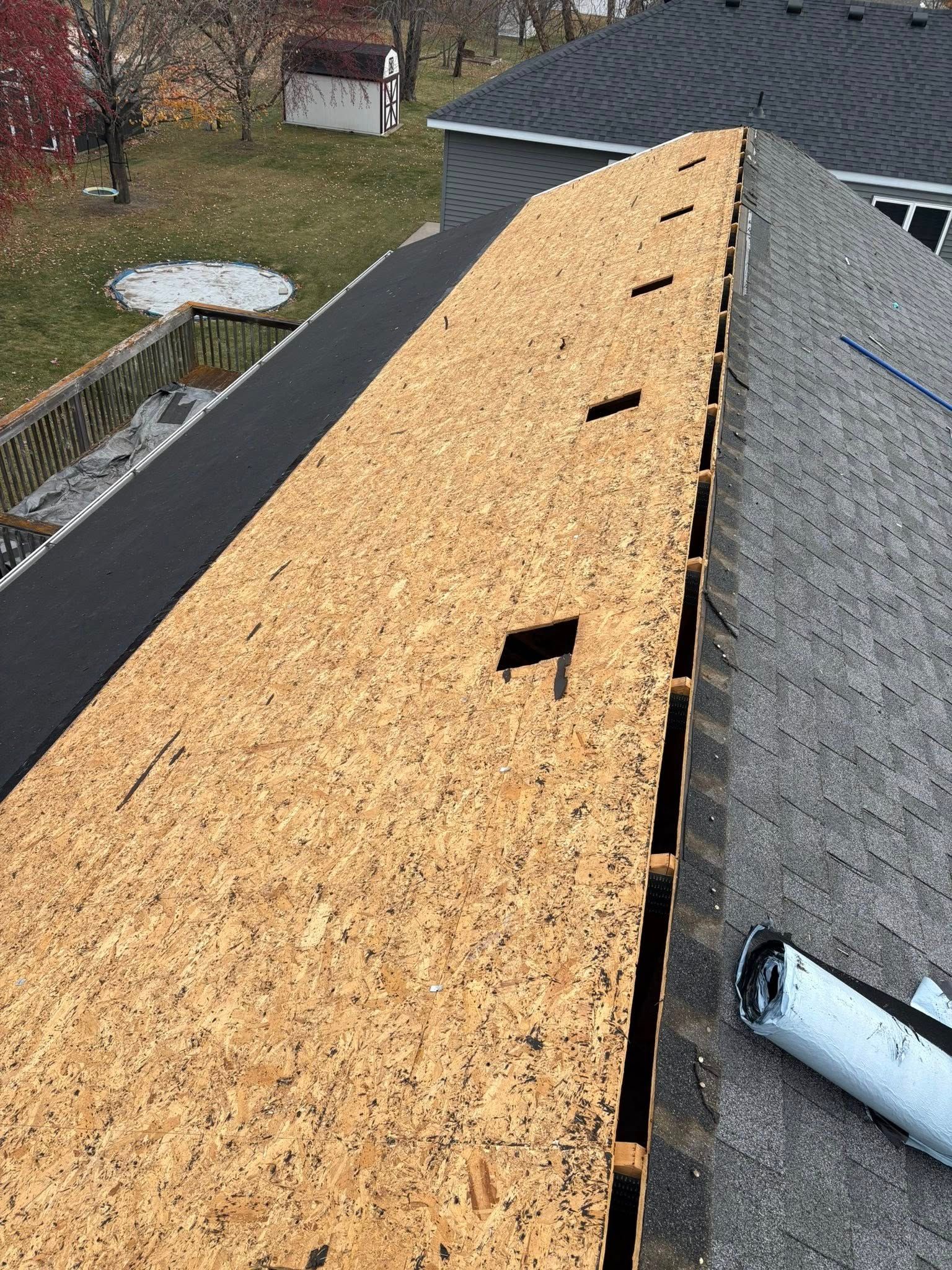 Roof under repair with exposed plywood underlayment beside gray shingles on a house roof