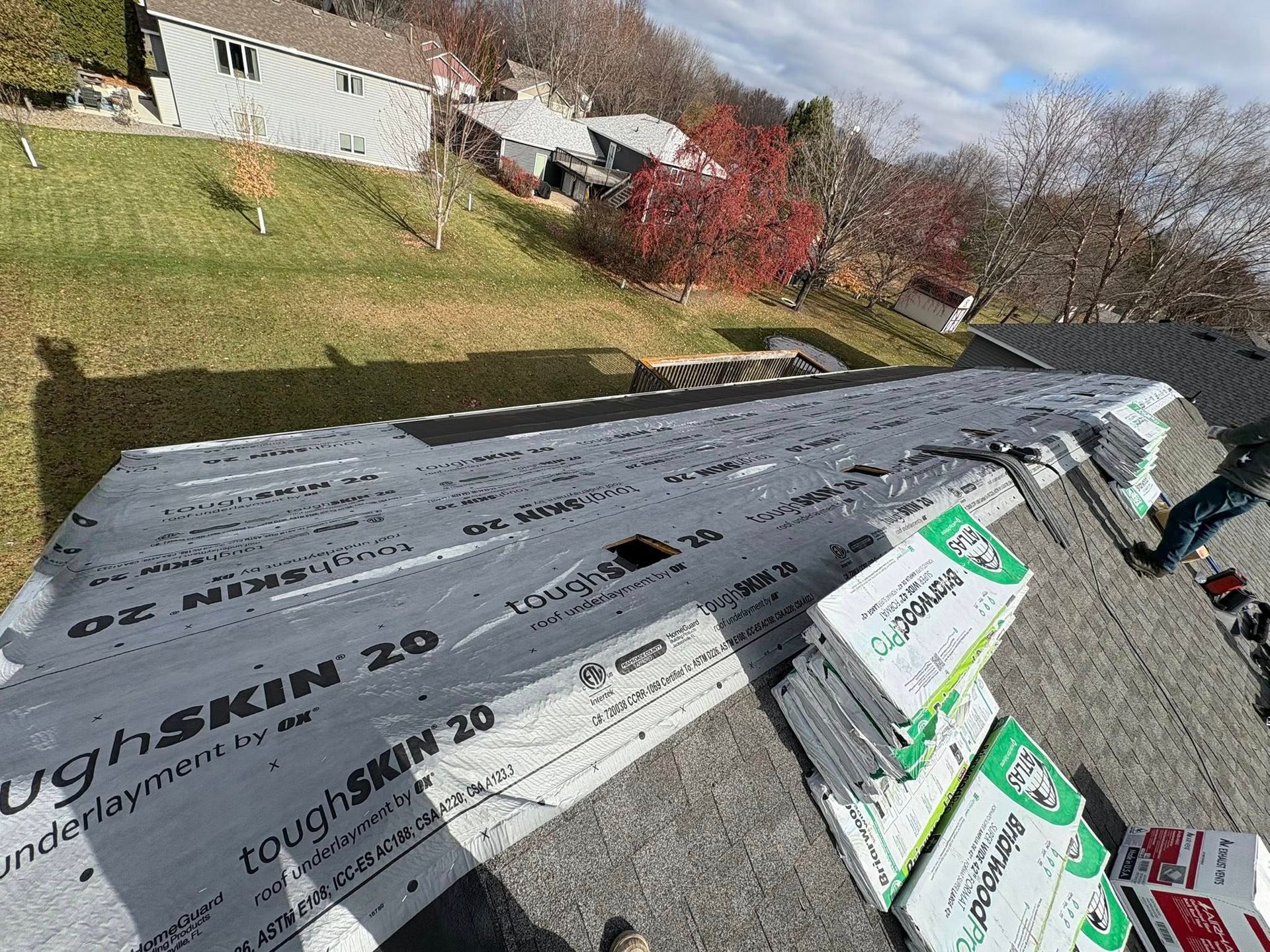 Roof under construction with gray underlayment and stacked shingles beside a grassy yard