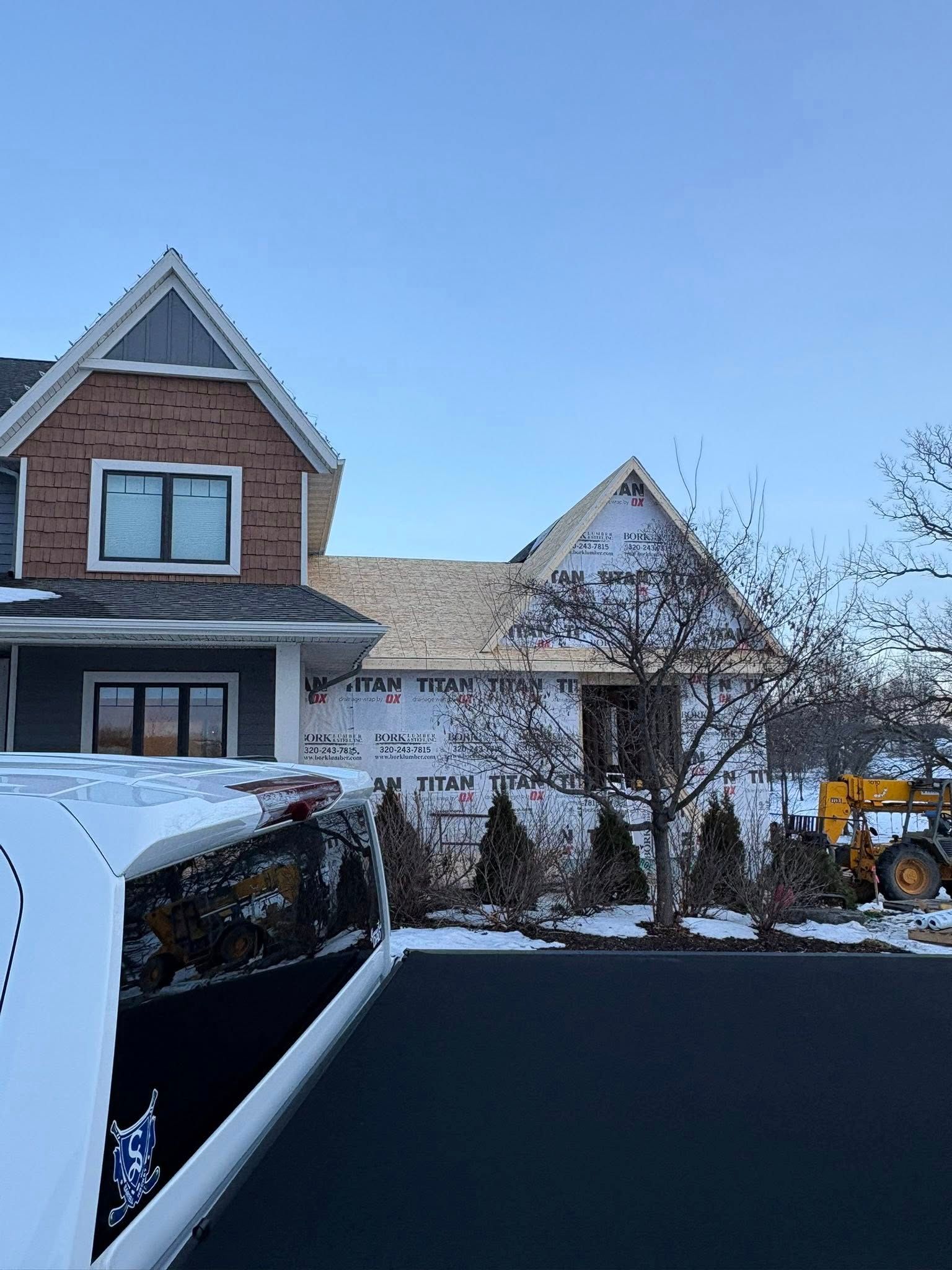 Snow-covered suburban house and driveway with a white van in the foreground and a yellow tractor at right.