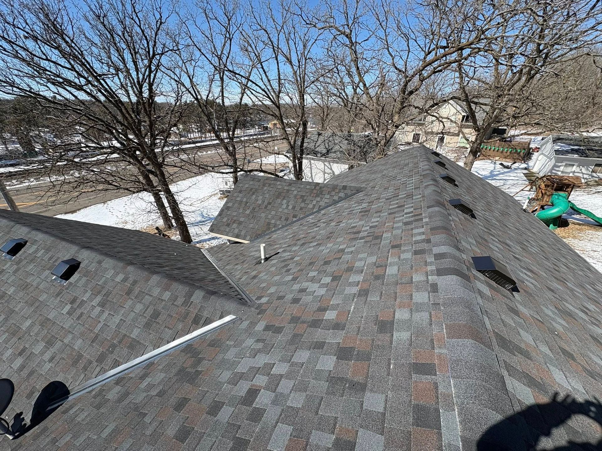 Snow-covered rooftop with gray shingles and bare trees in the background