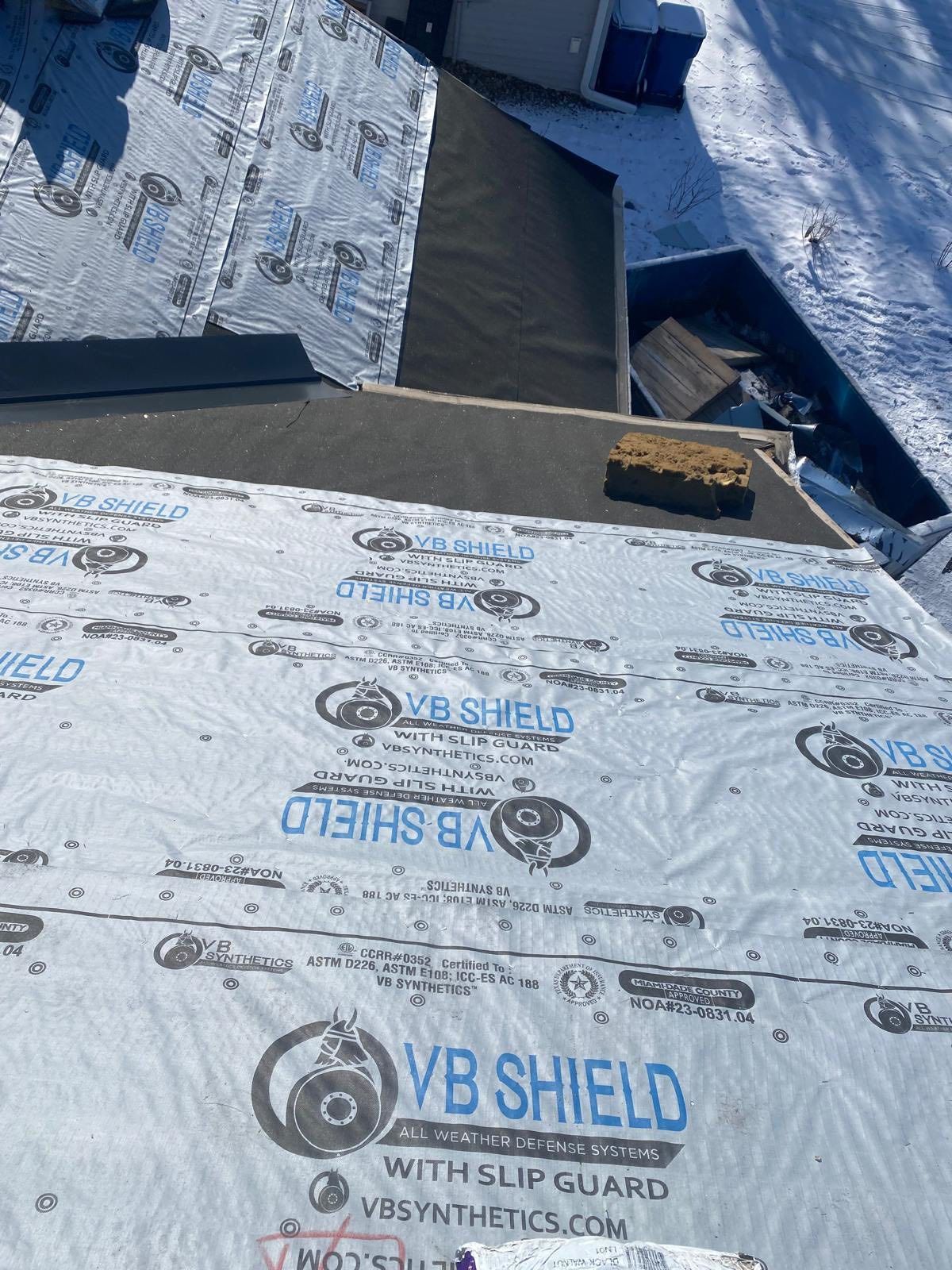 Blue roof underlayment with printed branding, torn seams, and a small yellow tool on the slope.