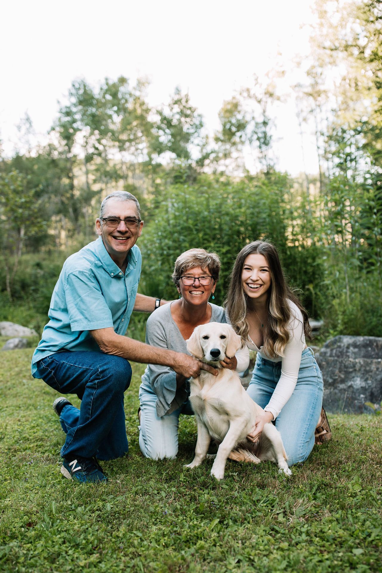 Family of three, a man, woman, and young woman, kneel with a golden retriever in a grassy outdoor setting.