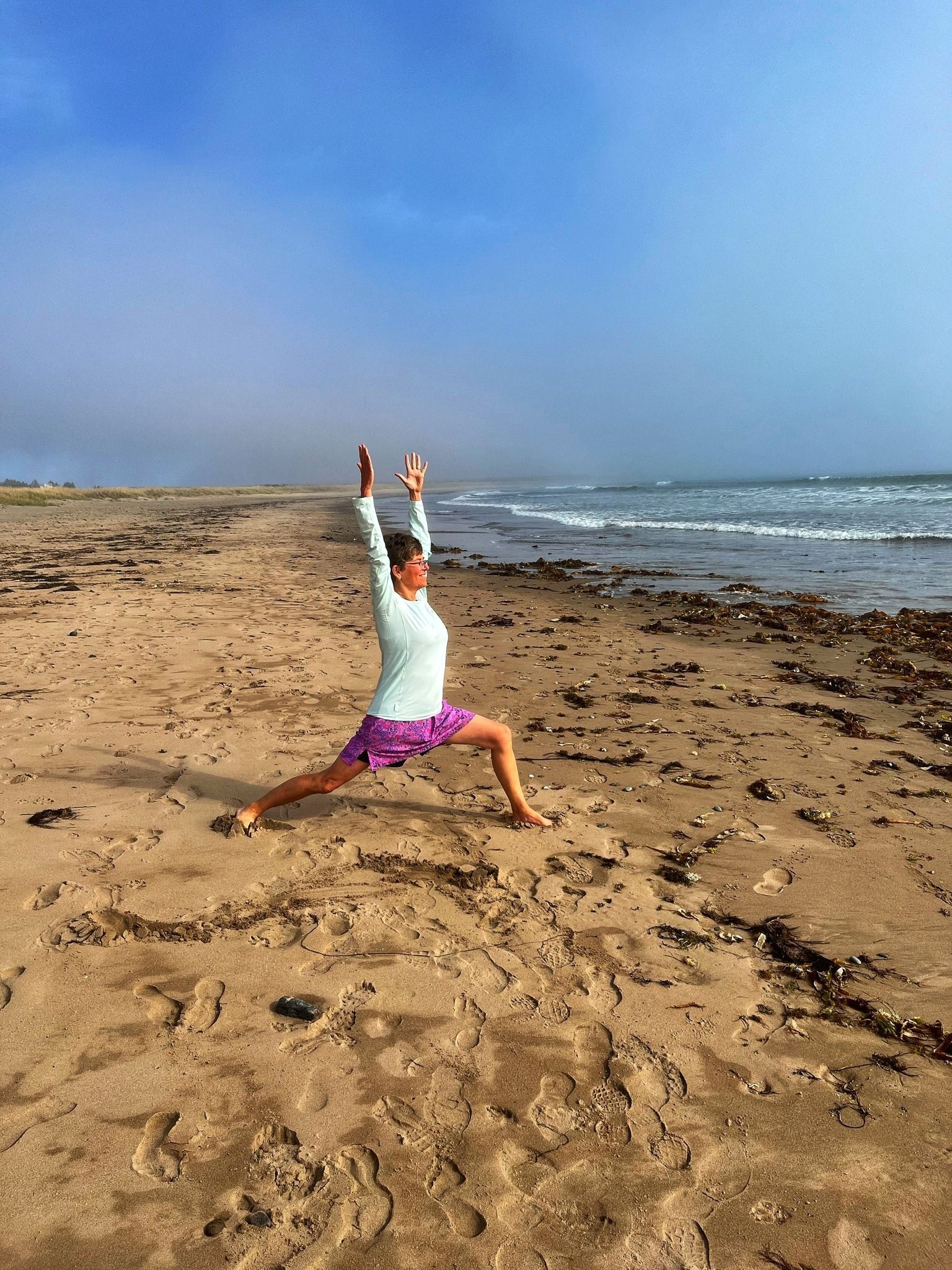 Person in a yoga pose on a sandy beach, arms raised, against a cloudy sky and ocean.
