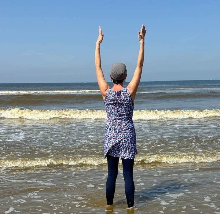 Woman in water with arms raised toward a blue sky over the ocean.