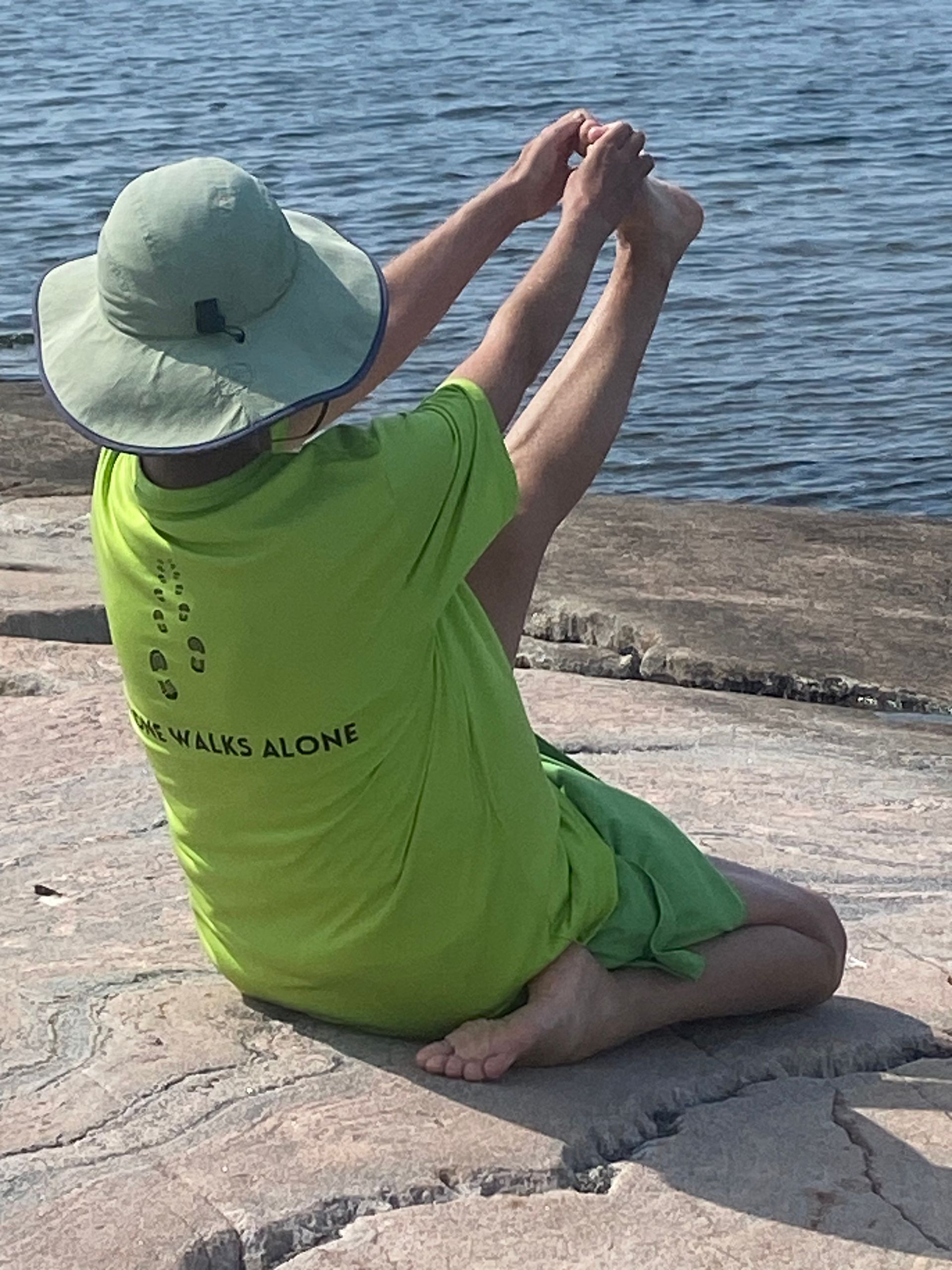 Person in green shirt and hat doing yoga on rocks by water, reaching for feet.