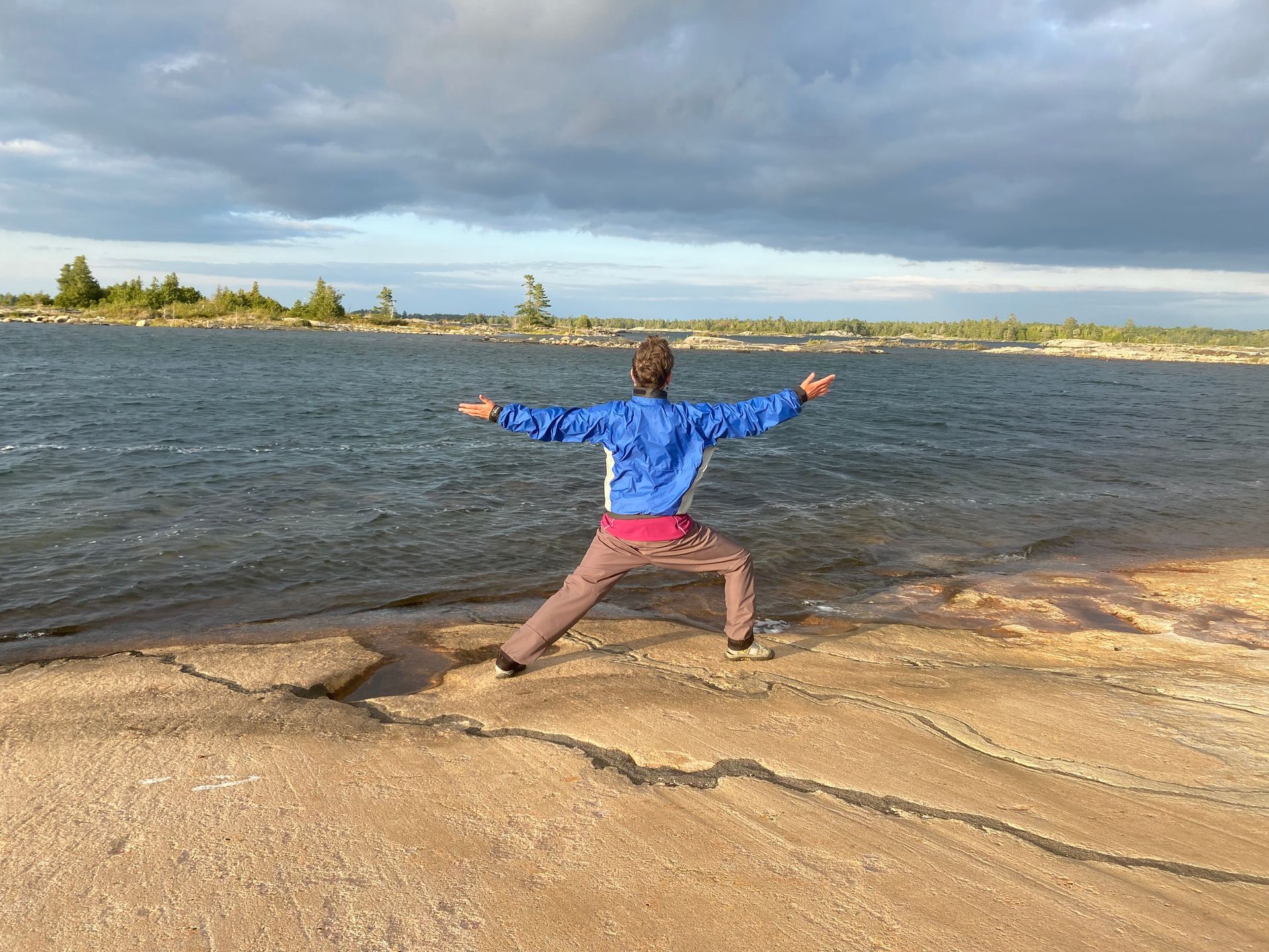 Person in blue jacket and brown pants in yoga pose on a rocky shoreline with a body of water in the background.