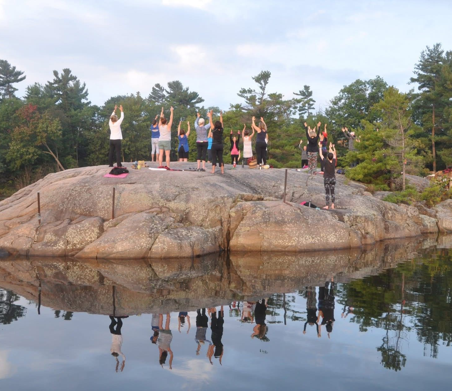 People doing yoga on a large rock, arms raised, reflected in calm water. Trees and sky in background.