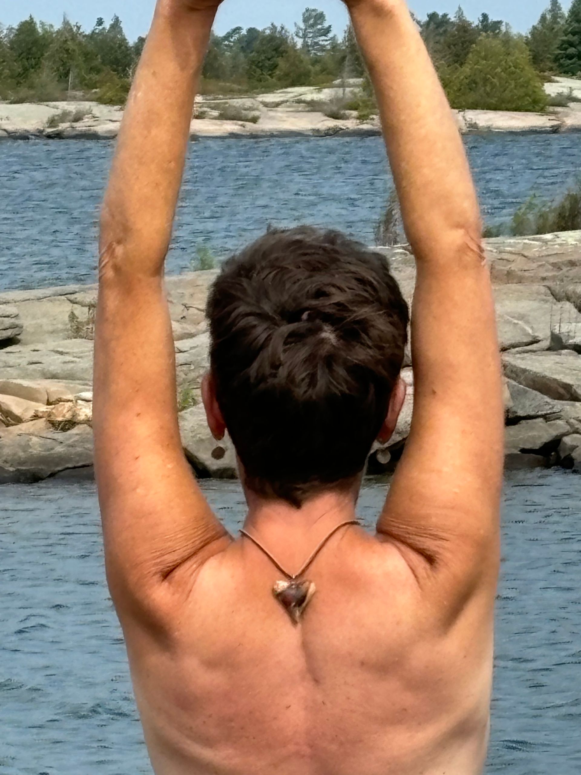 Person with arms raised overhead, back to the viewer, against a backdrop of water and rocks.