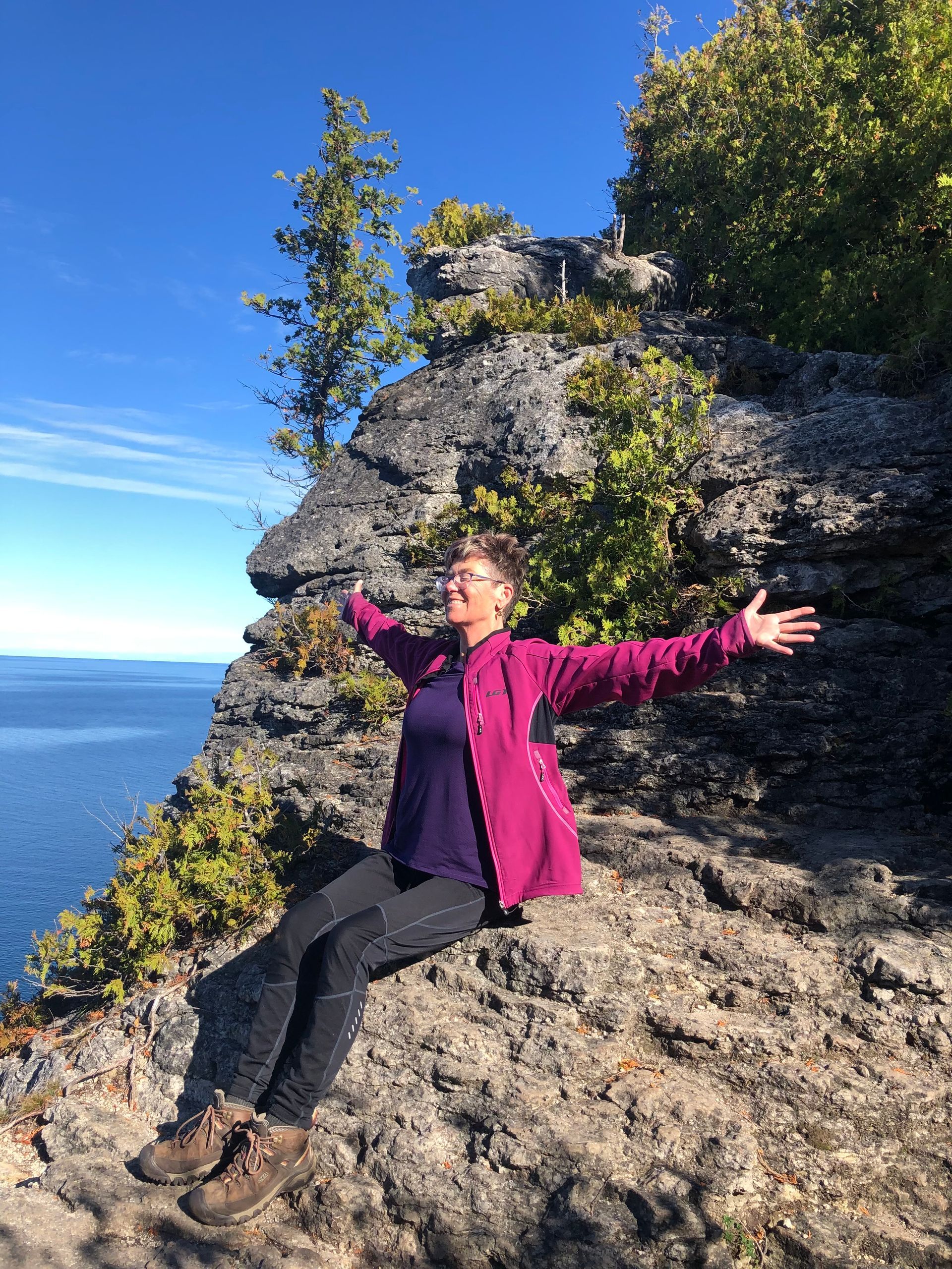 Woman with arms outstretched, smiles on cliff overlooking blue water and sky.