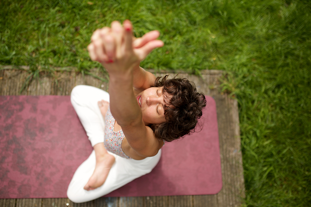 Woman in white pants and tank top doing yoga on a mat outdoors, arms raised, hands clasped, eyes closed.