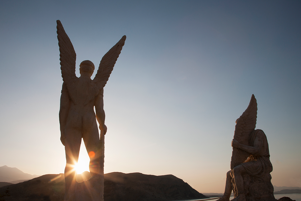 Statues of two figures with wings silhouetted against a sunrise, overlooking water.