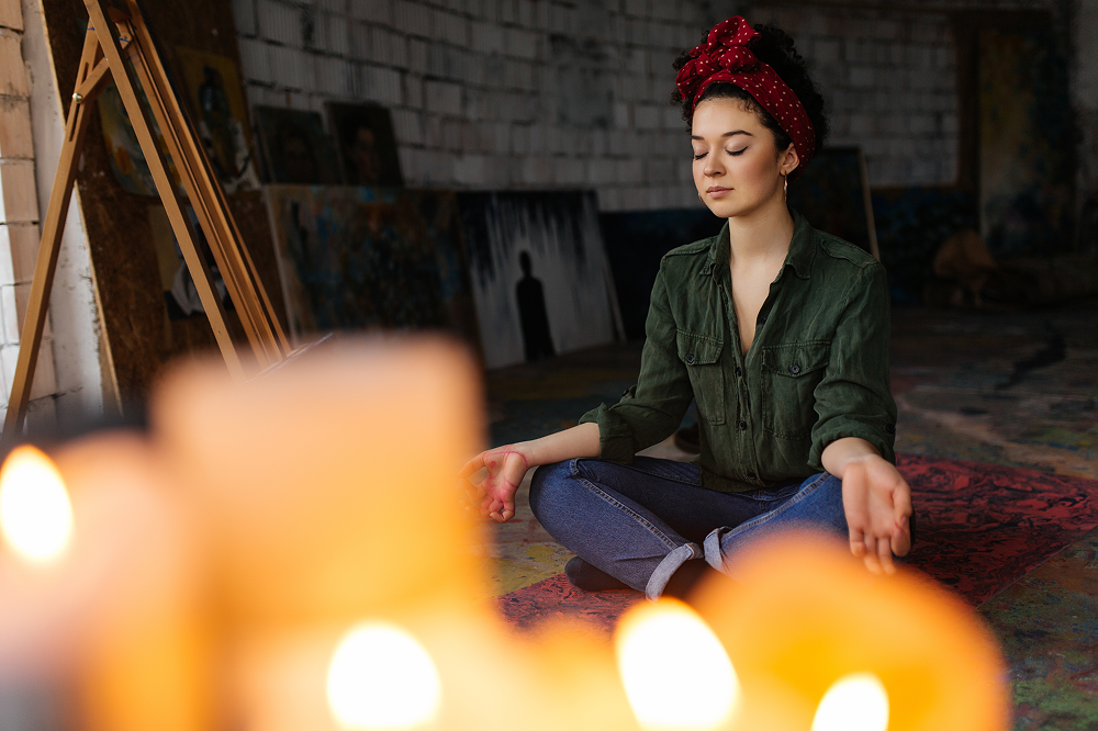 Woman in lotus pose meditating, surrounded by candles, in an art studio.