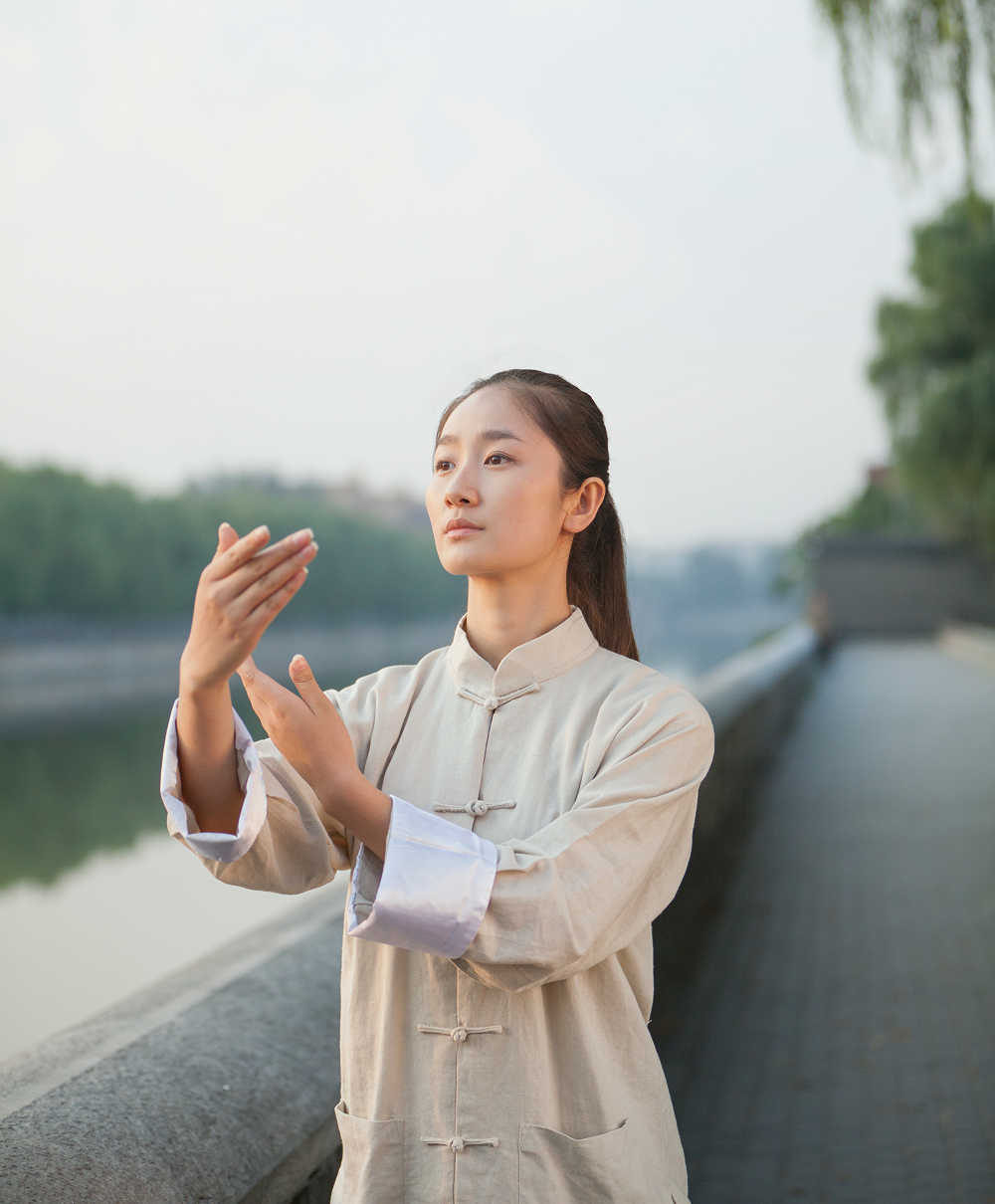 Woman in beige tunic practicing Tai Chi near a body of water, arms raised, focused expression.