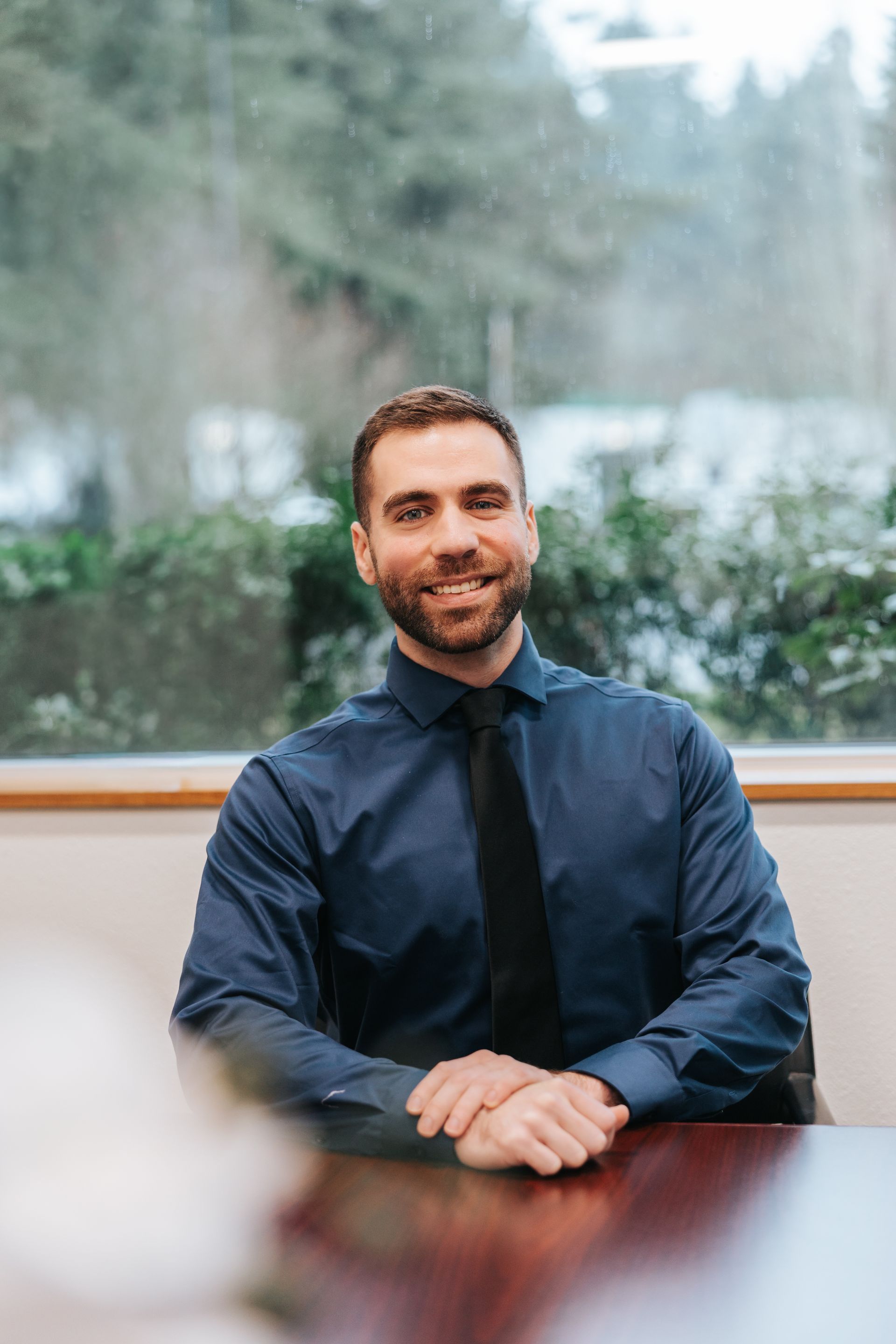 Man in light gray shirt, black tie, leaning against brick wall, smiling.