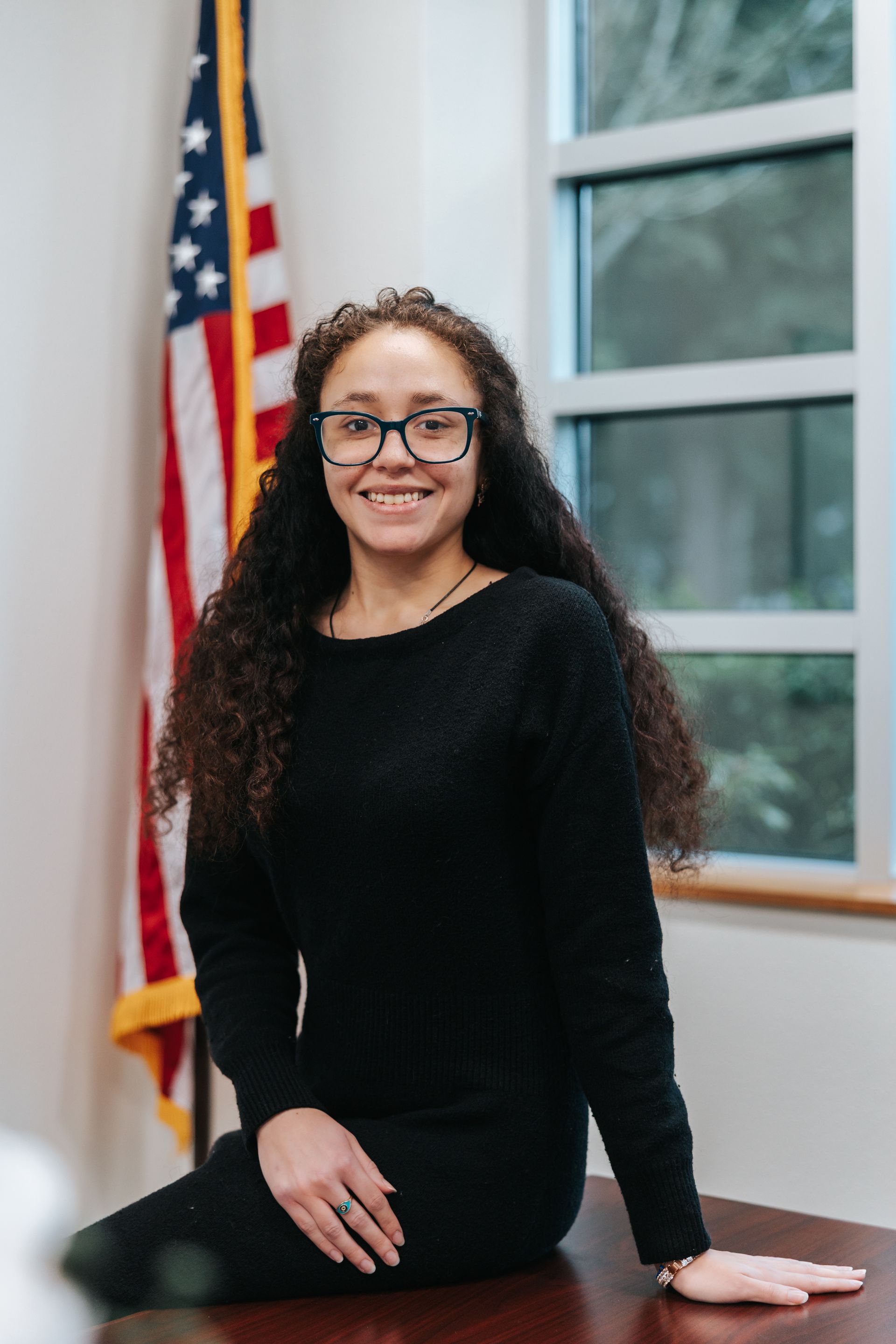 Woman in white dress and glasses smiles, arms crossed, by brick column.