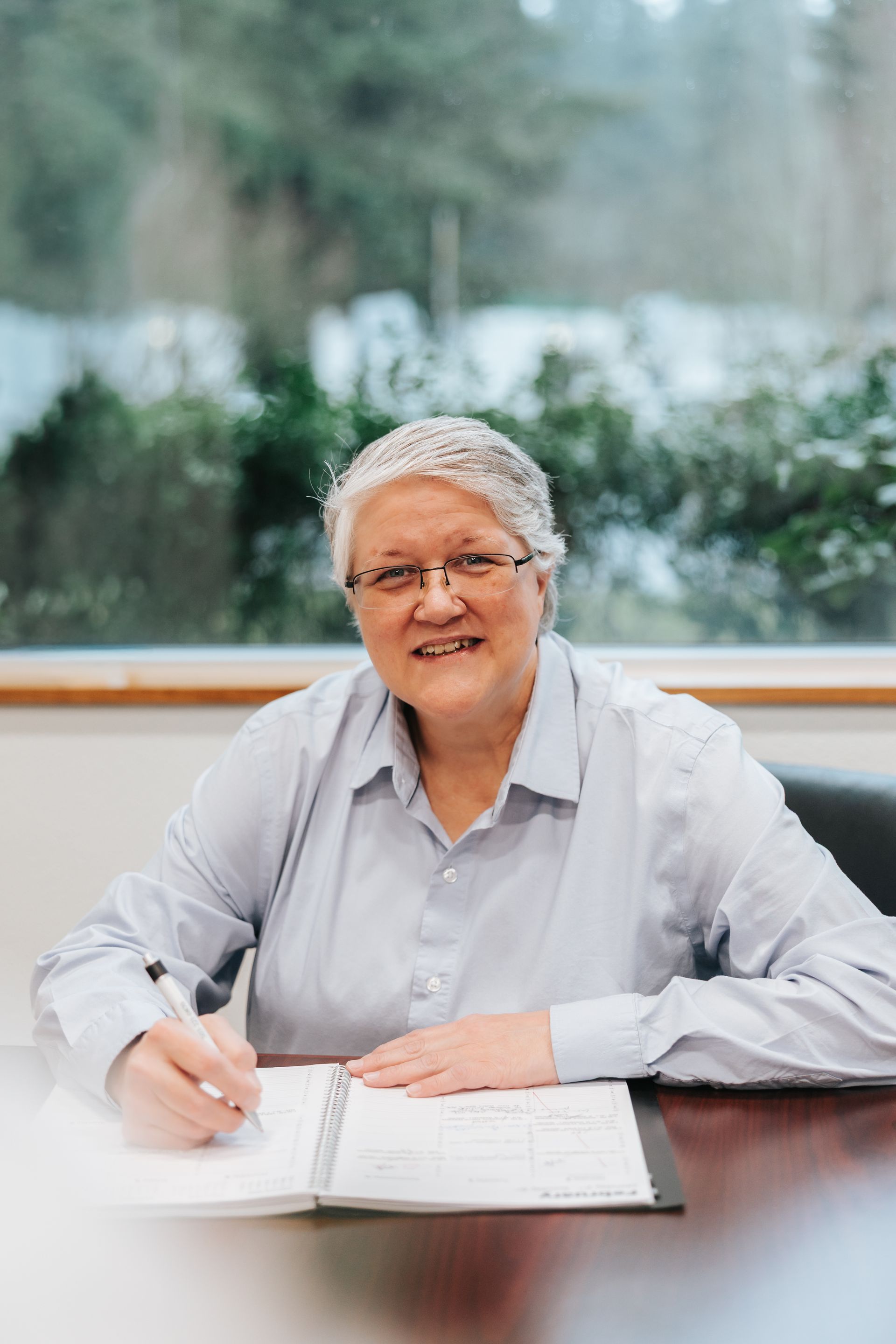 Person leaning against brick pillar, arms crossed, wearing a dark blue patterned shirt and jeans.