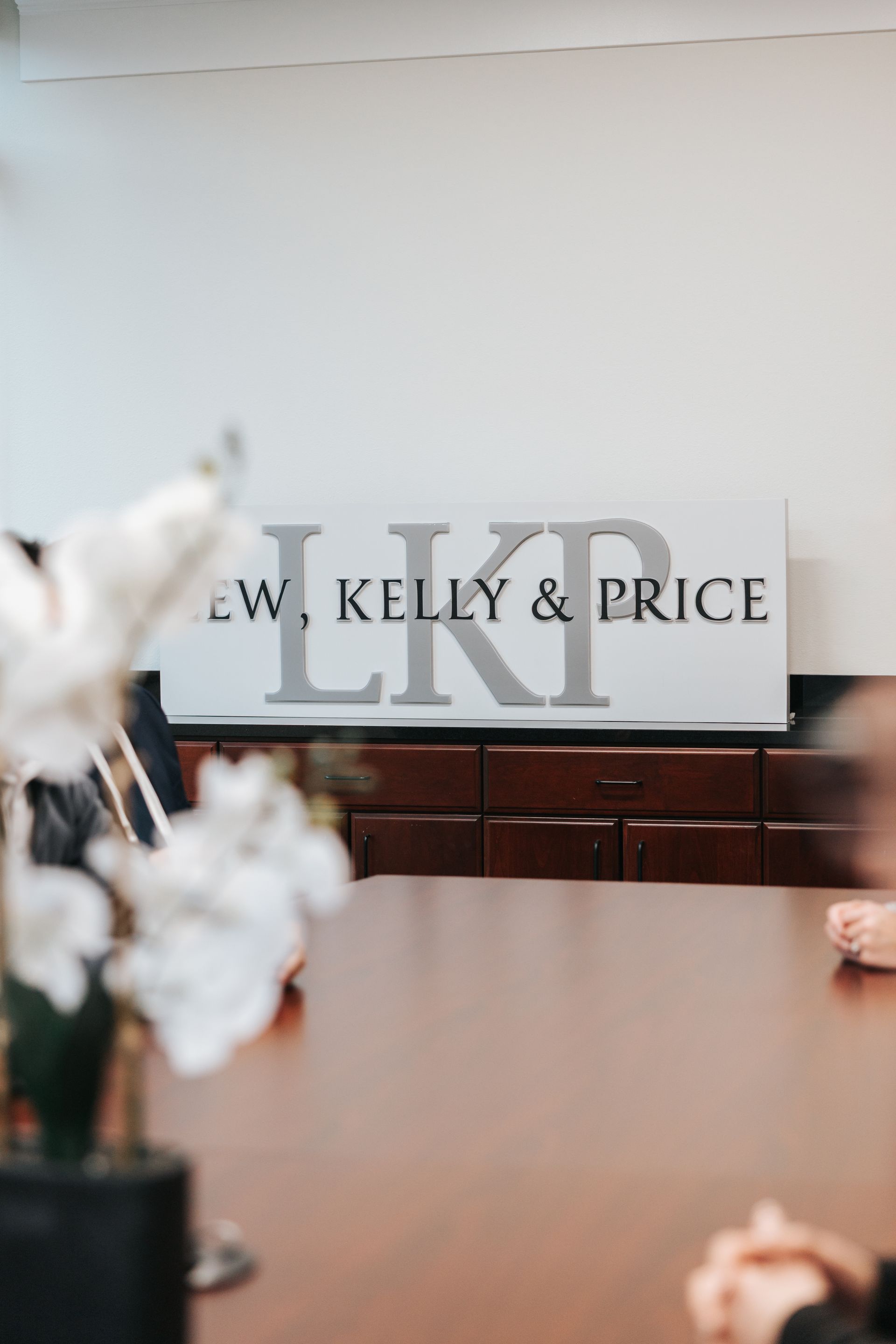 Woman with blonde hair smiling, wearing a floral dress, leaning against a brick column in a sunny hallway.